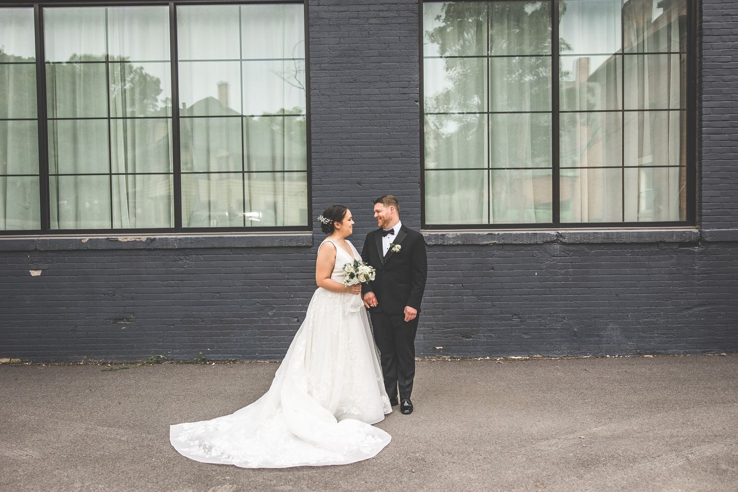 A couple in wedding attire poses together against a dark gray brick wall with large windows.