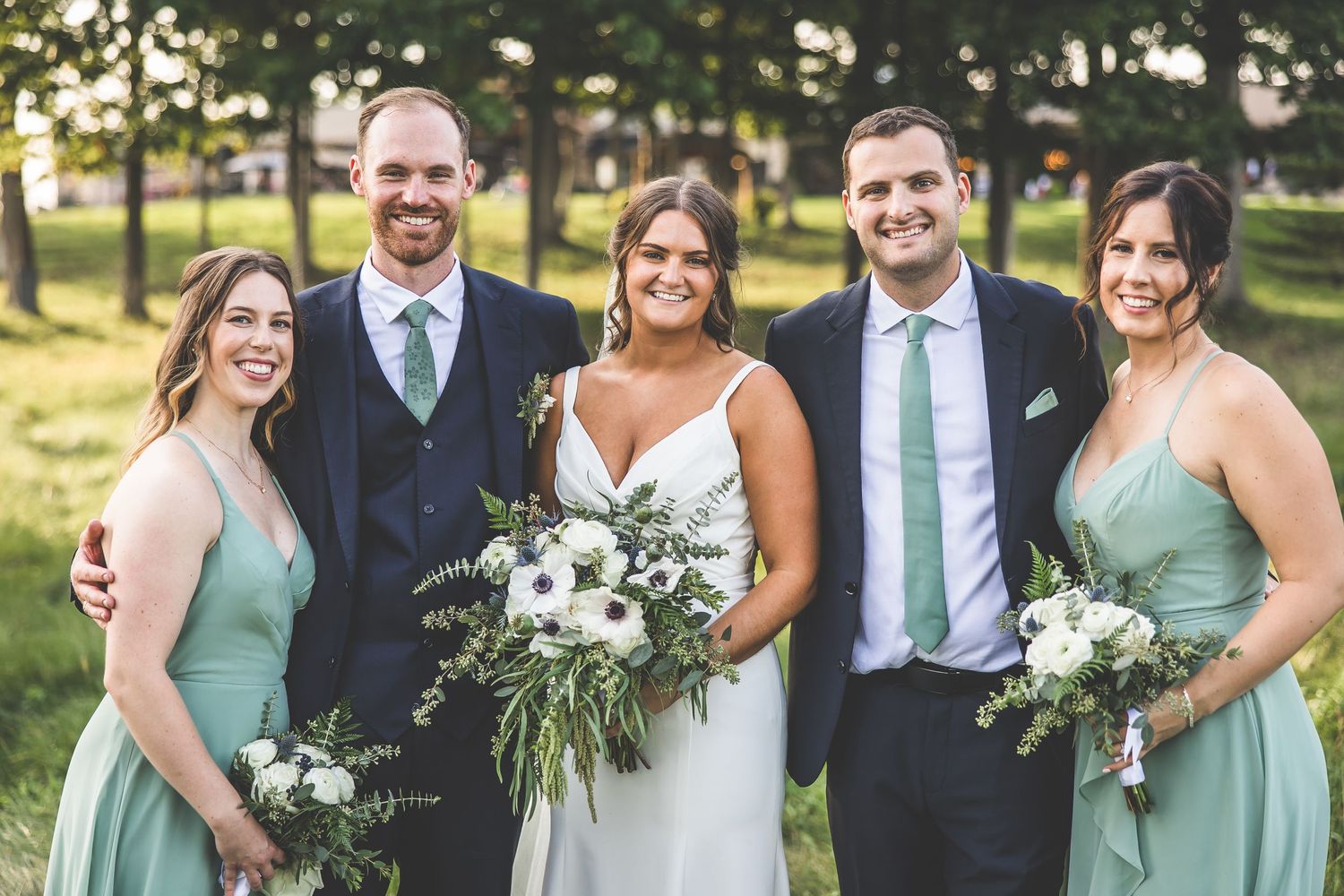 Wedding party in sage green dresses and navy suits pose together outdoors in a park setting.