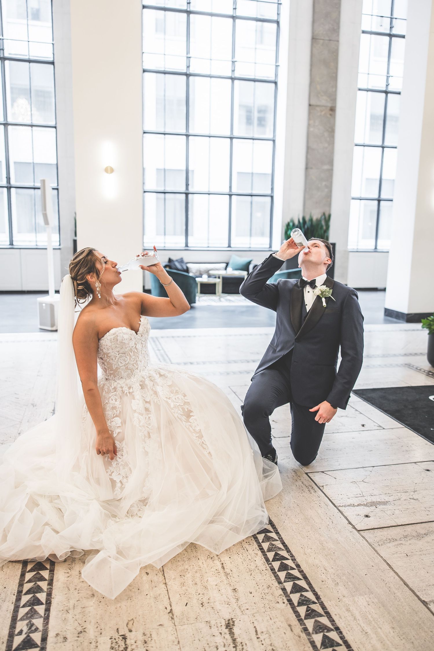 Playful wedding couple takes fun photos while lying on decorative floor in bright venue.