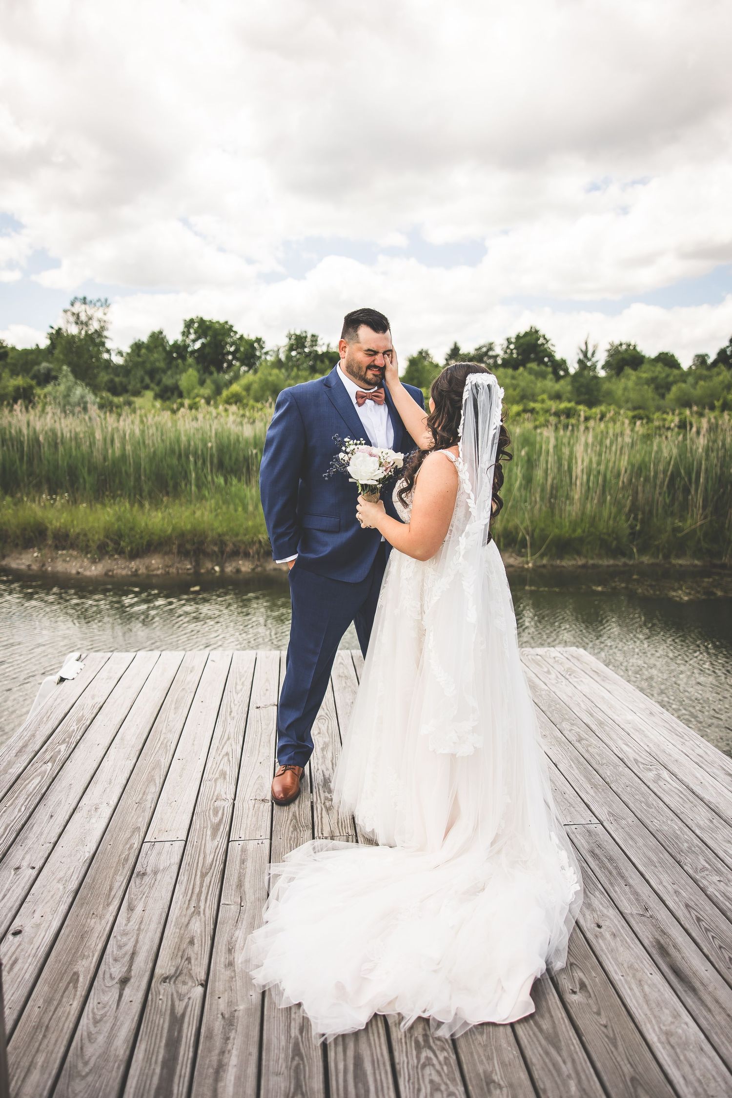 Wedding couple embraces on a wooden dock with scenic lake and greenery in the background.