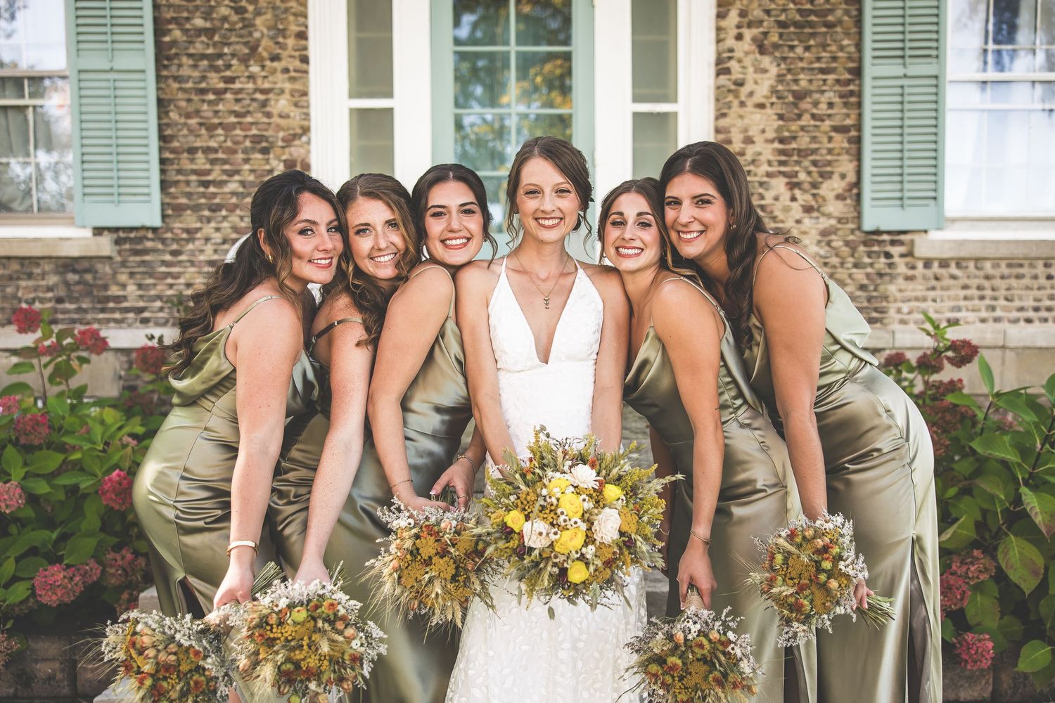 Bridesmaids in sage green dresses pose with bouquets alongside bride in white dress against brick building with green shutters.