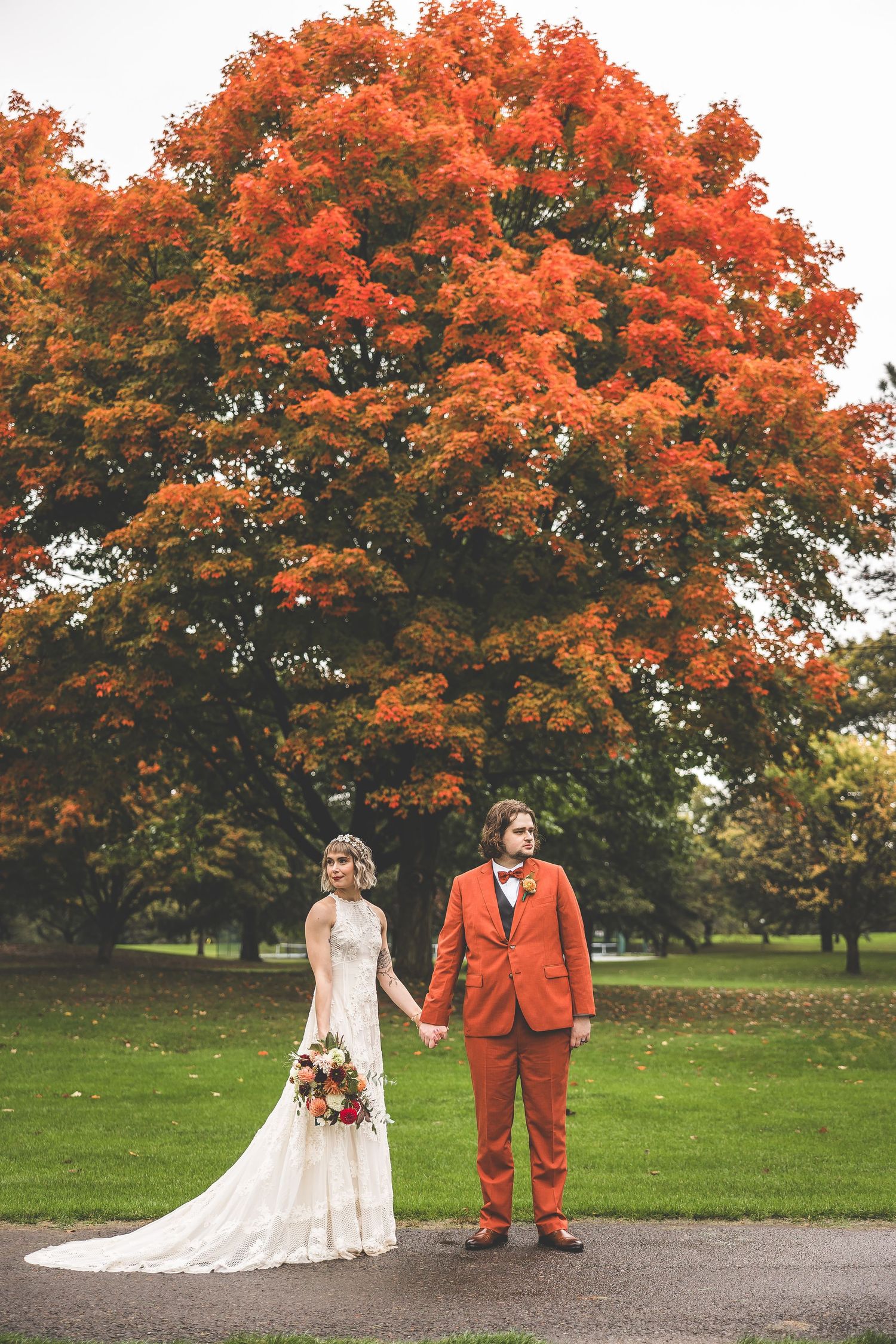 A couple in wedding attire poses under vibrant orange autumn trees on a paved path.