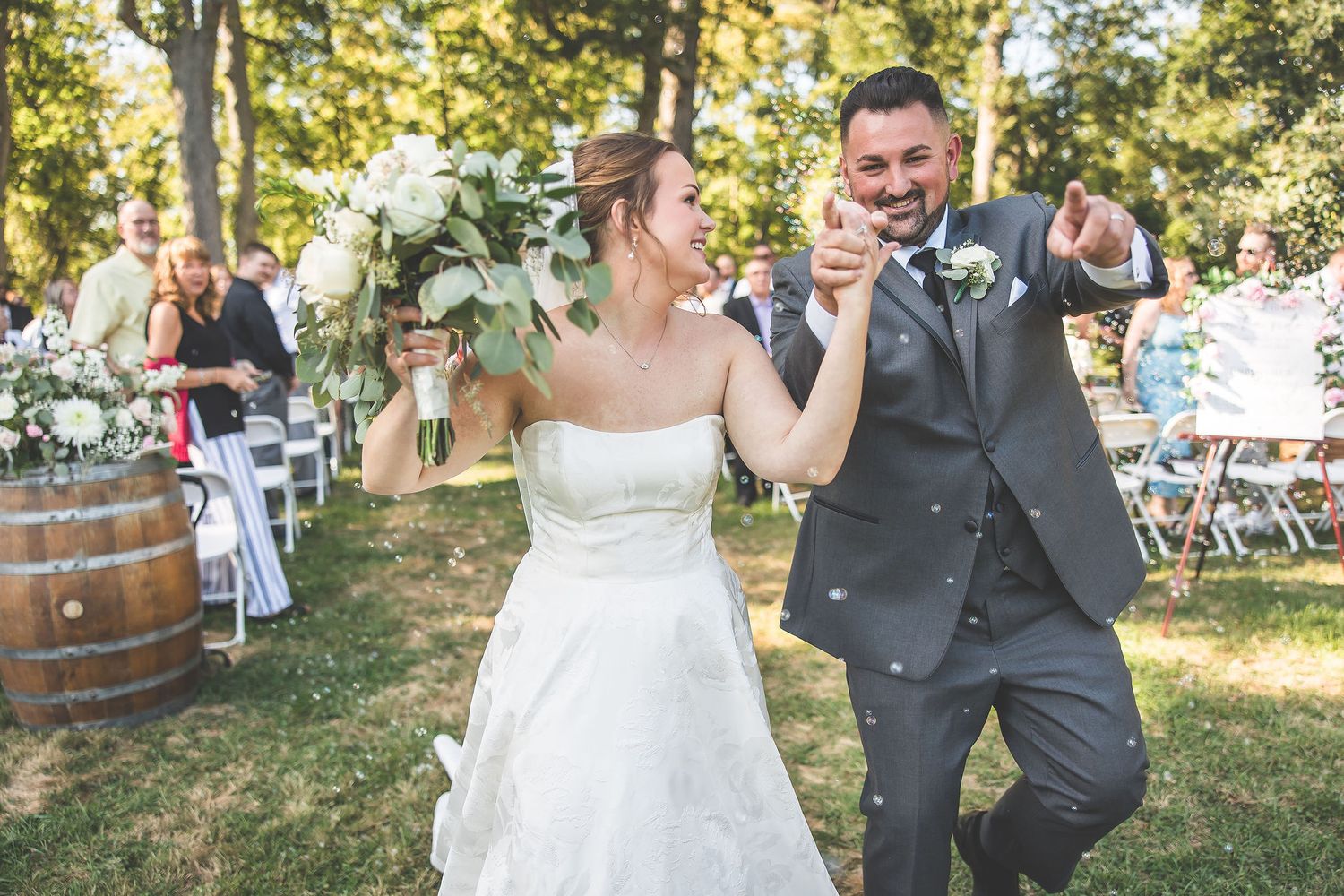 Wedding party celebrates during a sunny outdoor ceremony surrounded by tall trees.