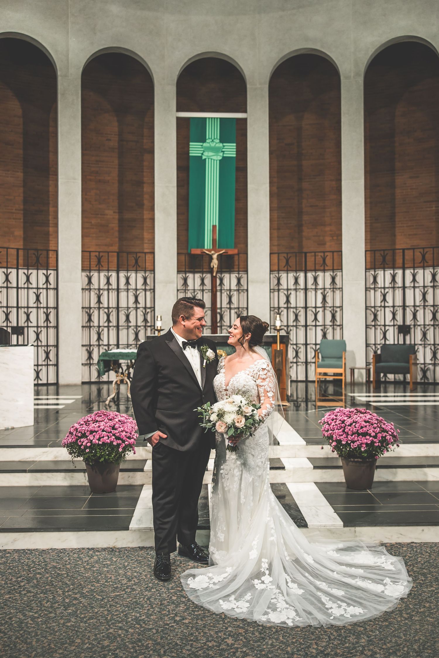 Elegant wedding couple poses in front of arched colonnade with pink floral arrangements and turquoise cross.