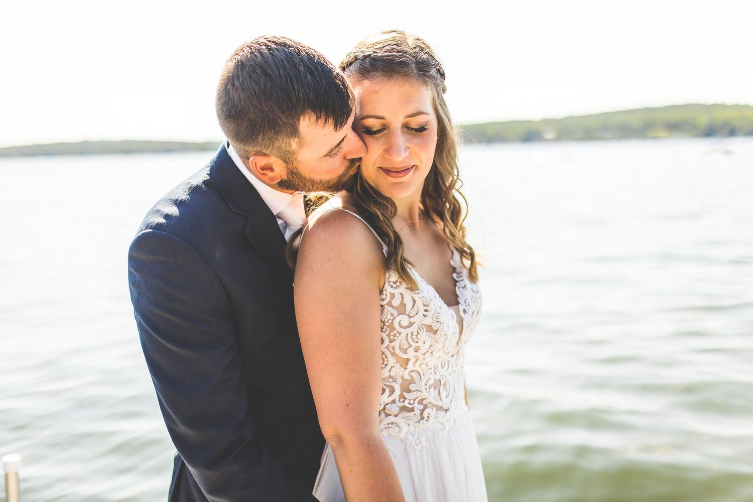 Romantic moment between newlyweds captured against lakefront backdrop.