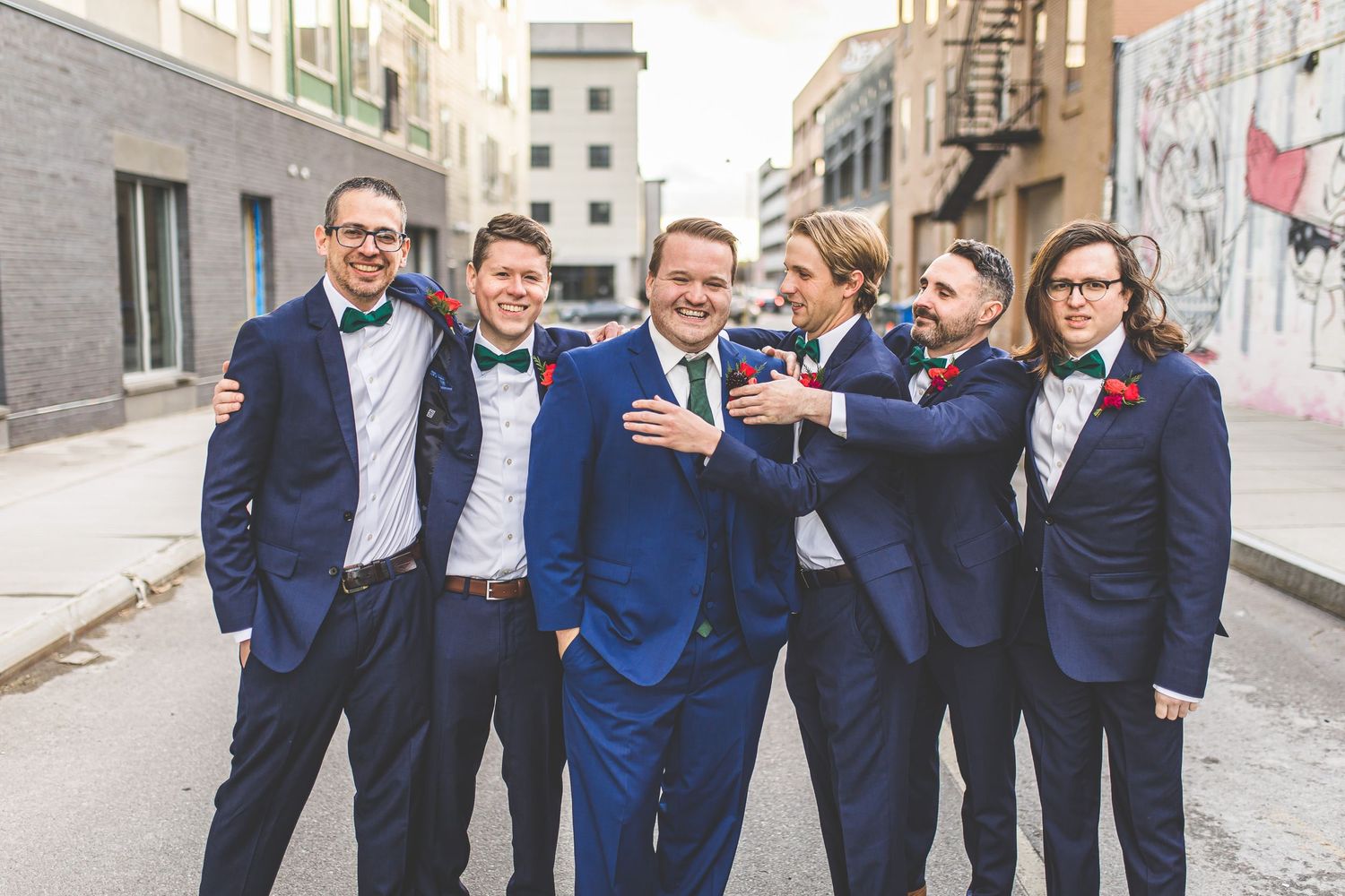 Wedding party laughing and posing together on city street in navy suits and green dresses.