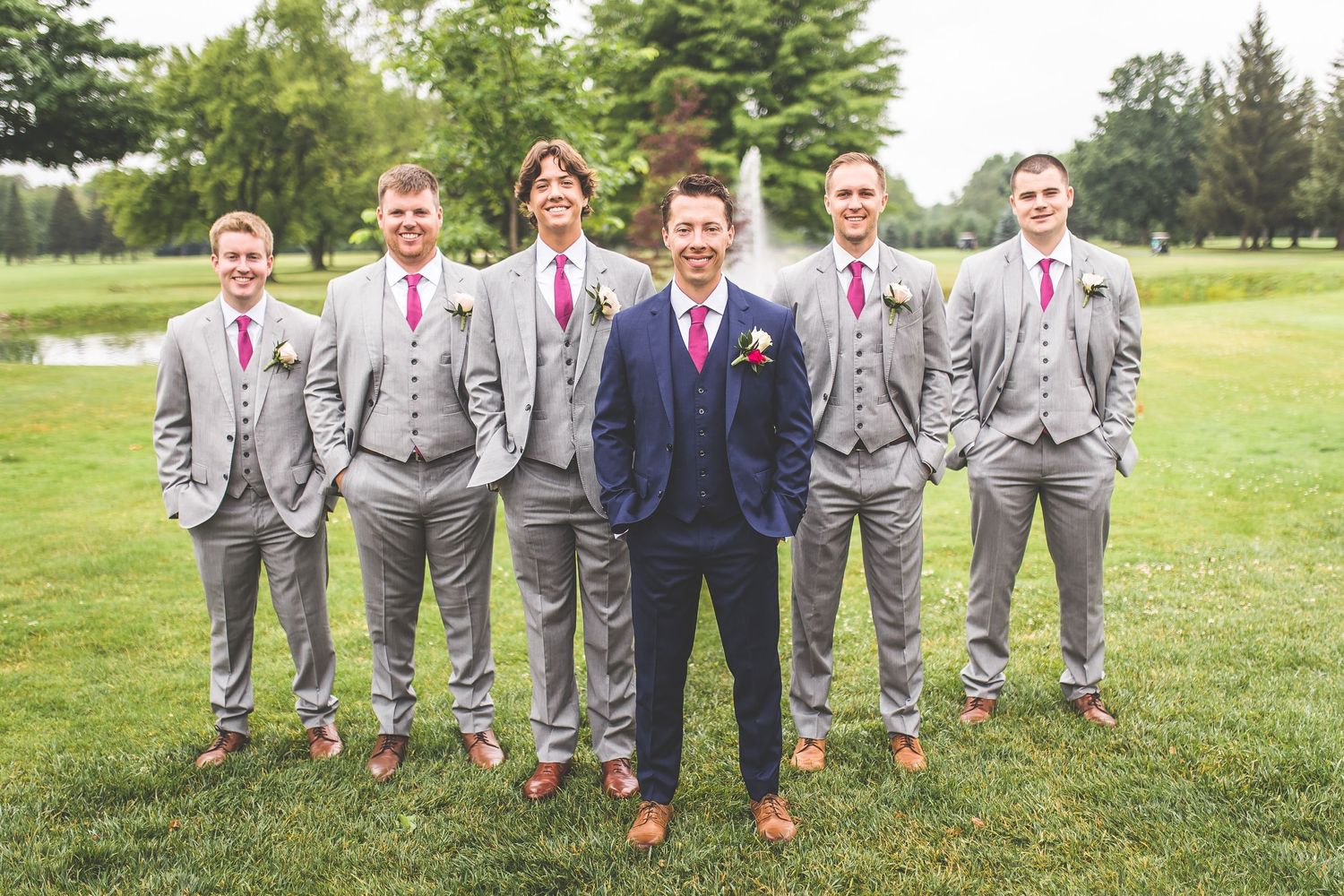 Groomsmen in gray suits with pink accents pose together on grass for wedding party photos.