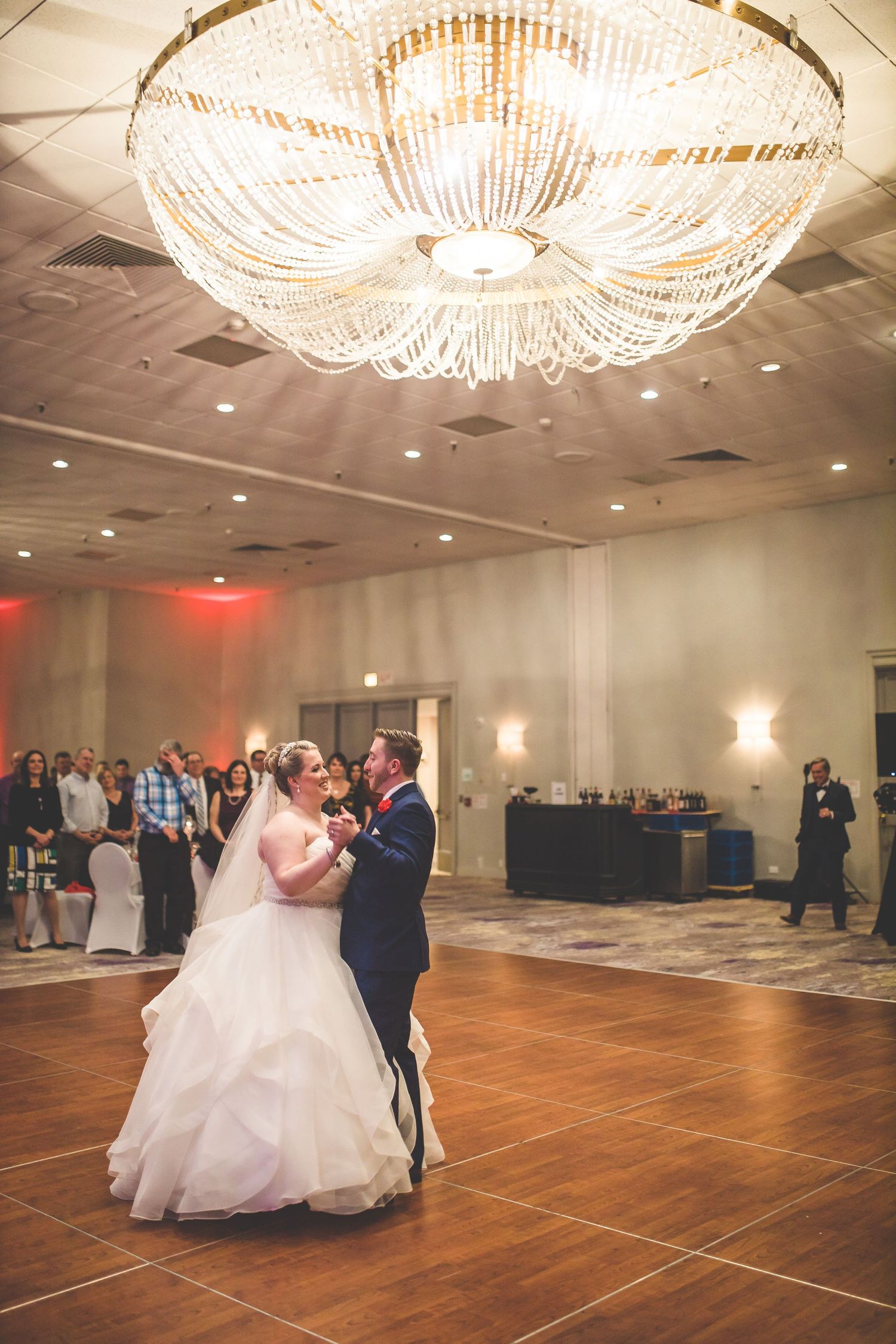 Couple dances beneath an ornate crystal chandelier on wooden ballroom floor.