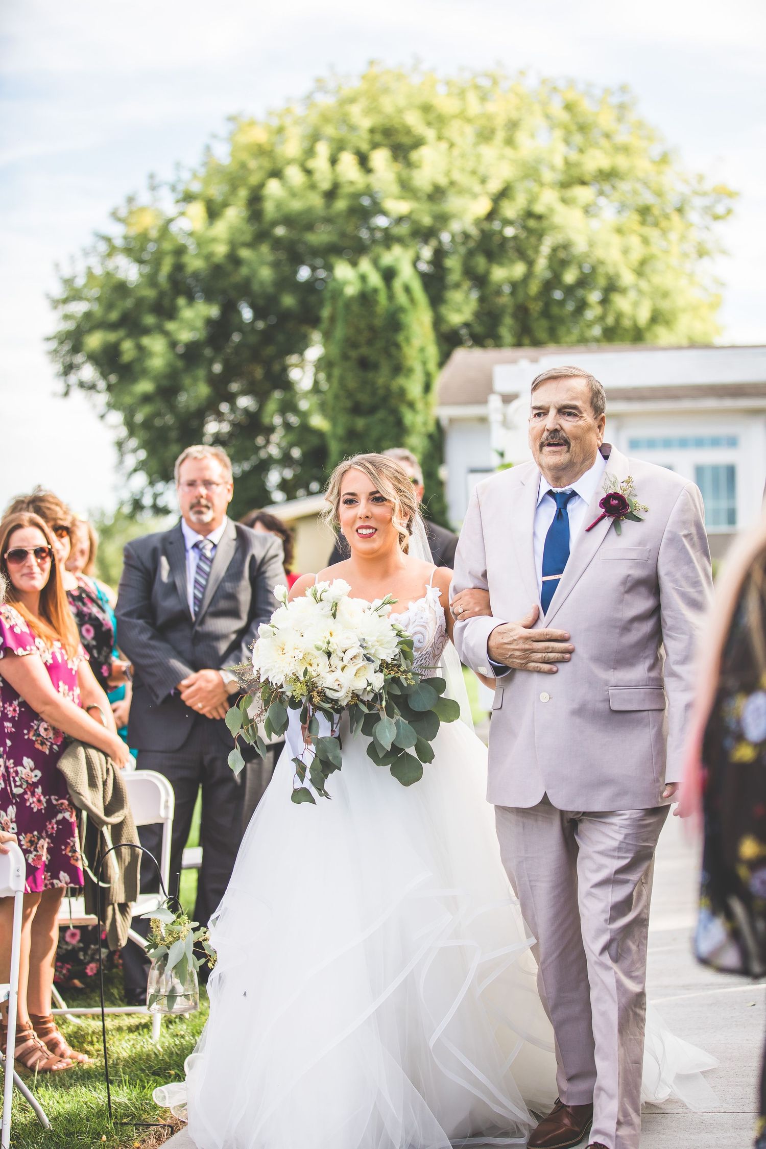 Outdoor wedding ceremony processional with guests in summer garden setting.