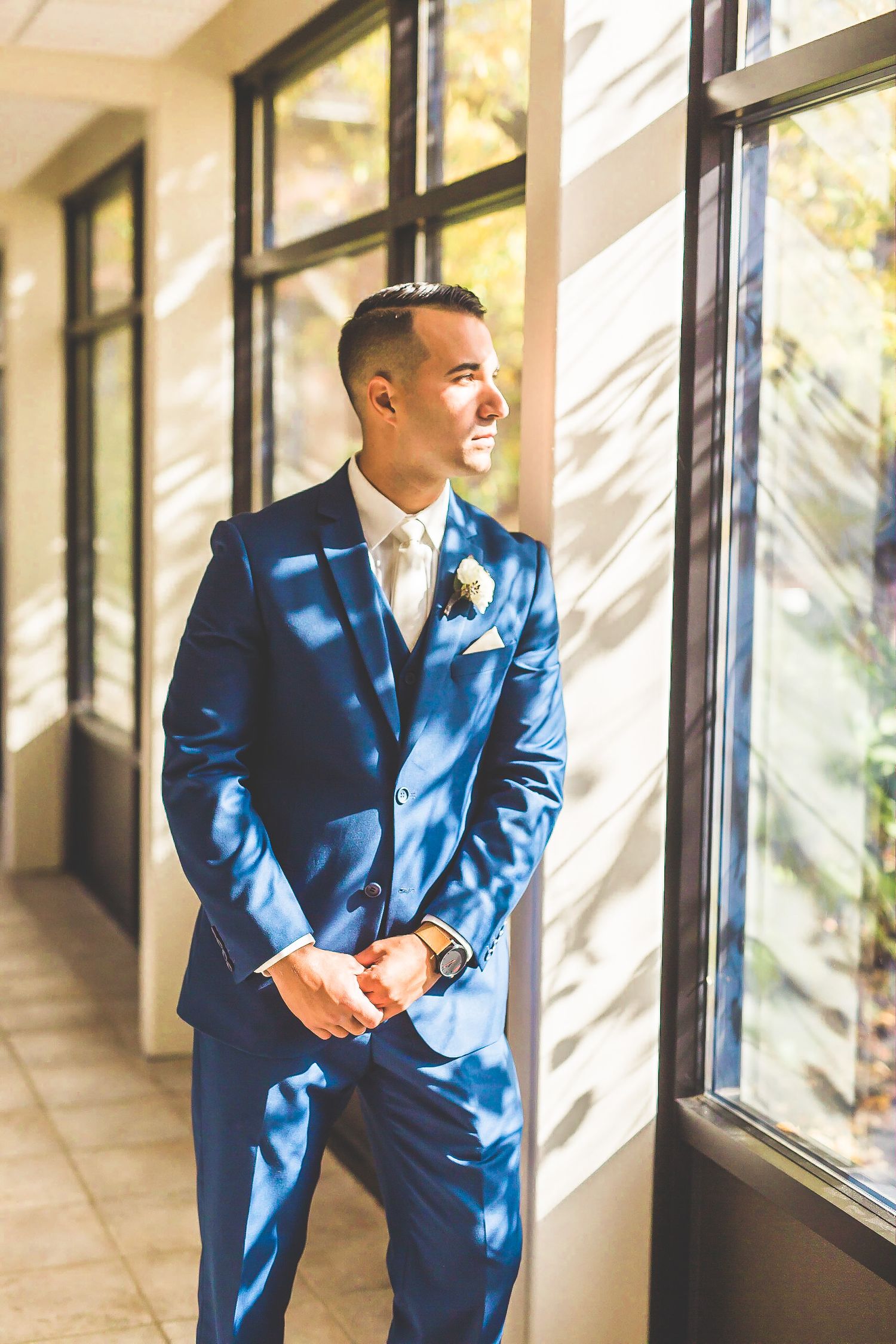 Groom in blue suit standing in sunlit hallway with window shadows.
