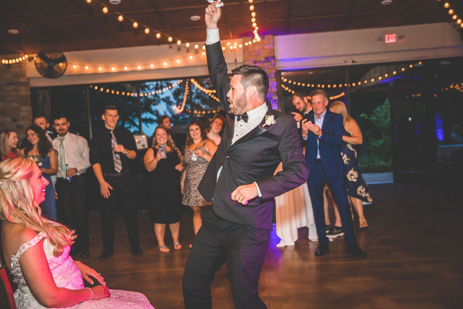 Wedding guests dance and celebrate under string lights at an indoor reception venue.