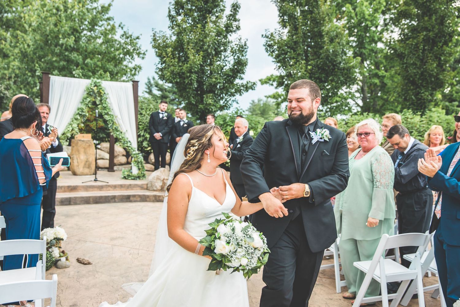 Outdoor wedding ceremony with white chairs and floral arrangements under trees.