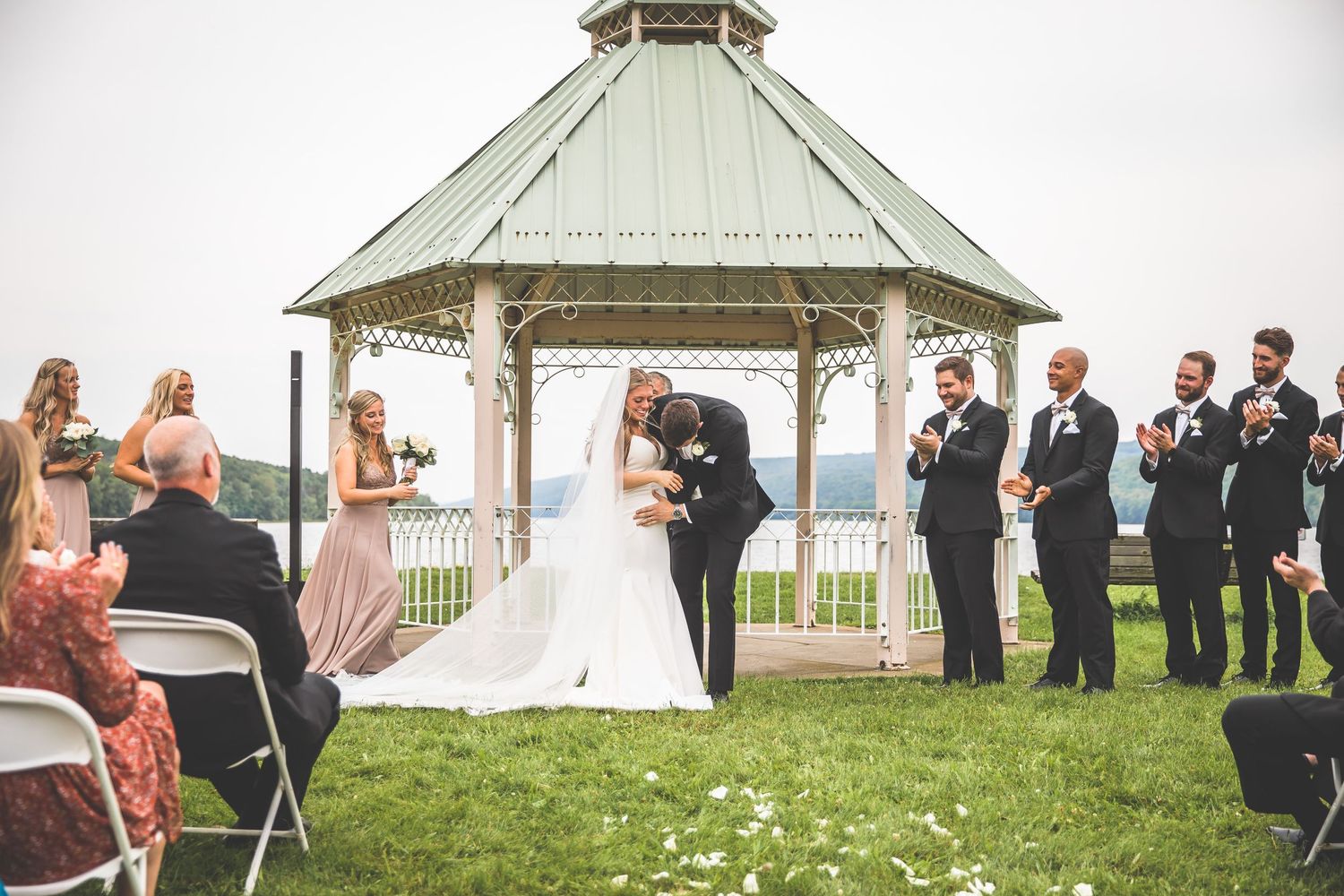 Outdoor wedding ceremony arrangement with guests seated under cloudy skies by a lakeside gazebo.