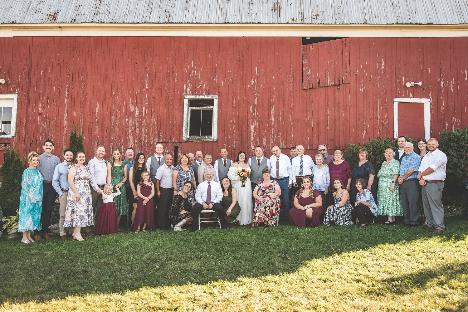 Panoramic group shot of wedding attendees posed in front of a classic red barn.