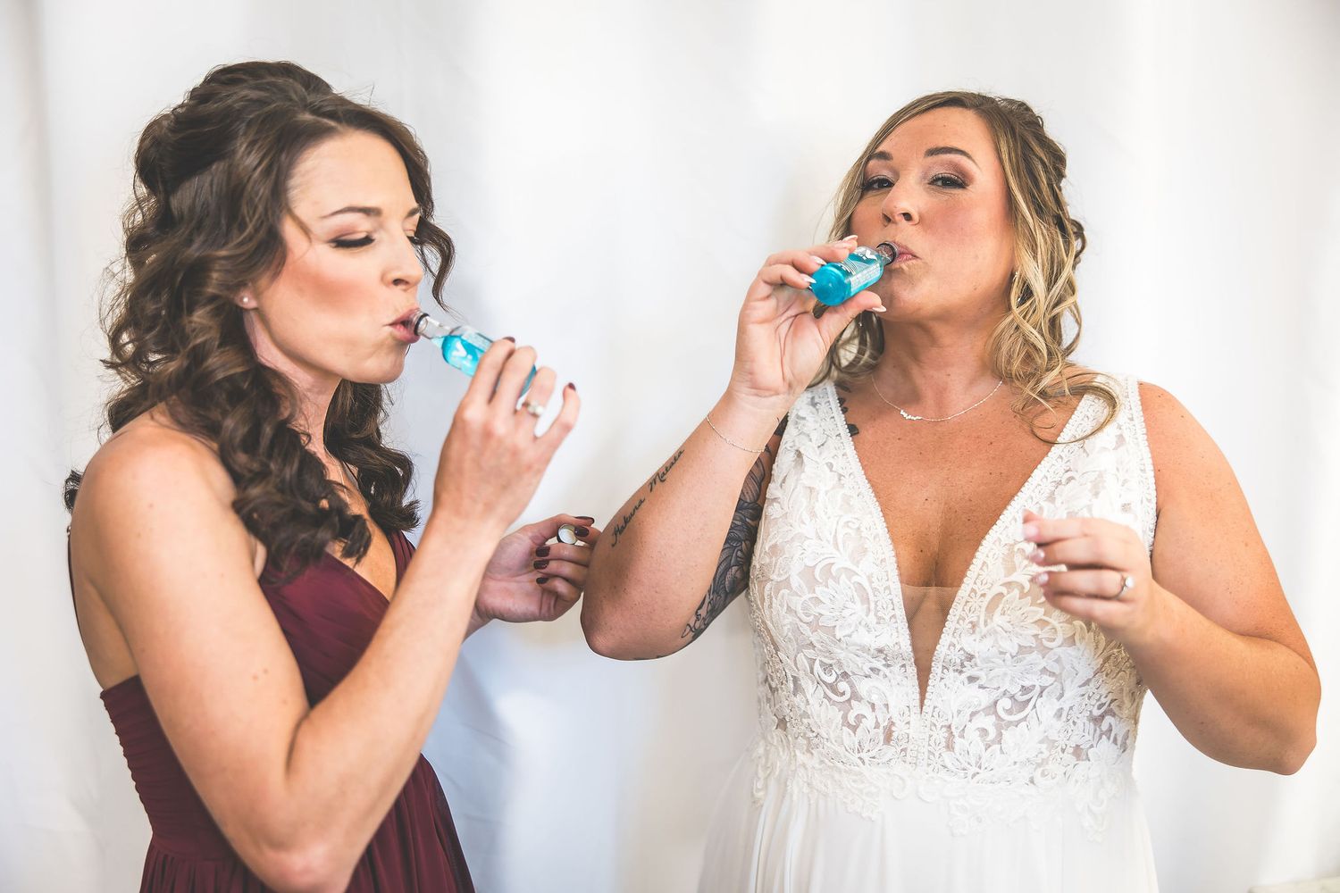 Bridesmaids in burgundy dresses and bride in white share a celebratory toast together in a bright room.