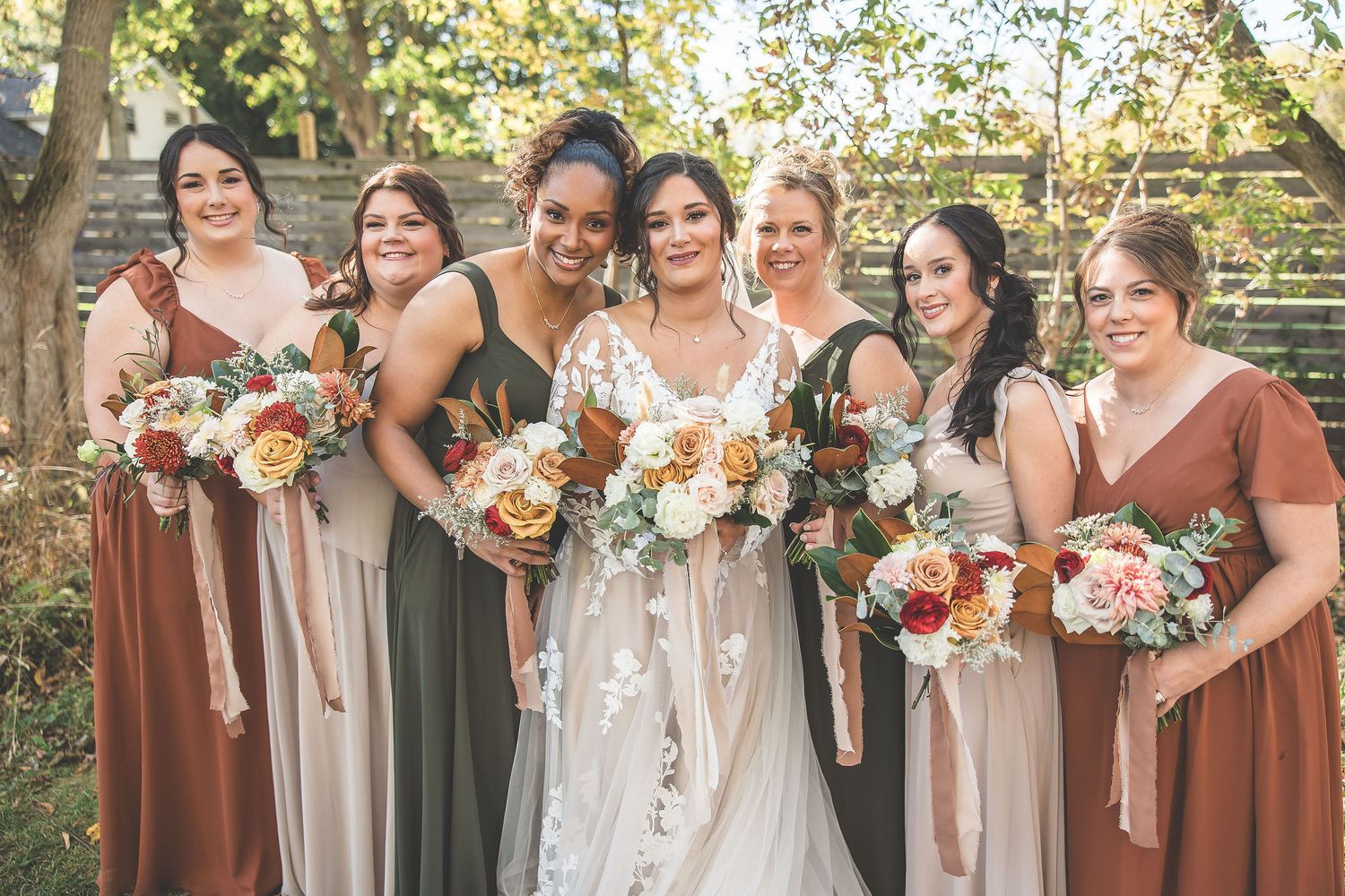 Bridal party poses together outdoors wearing burgundy and neutral dresses.