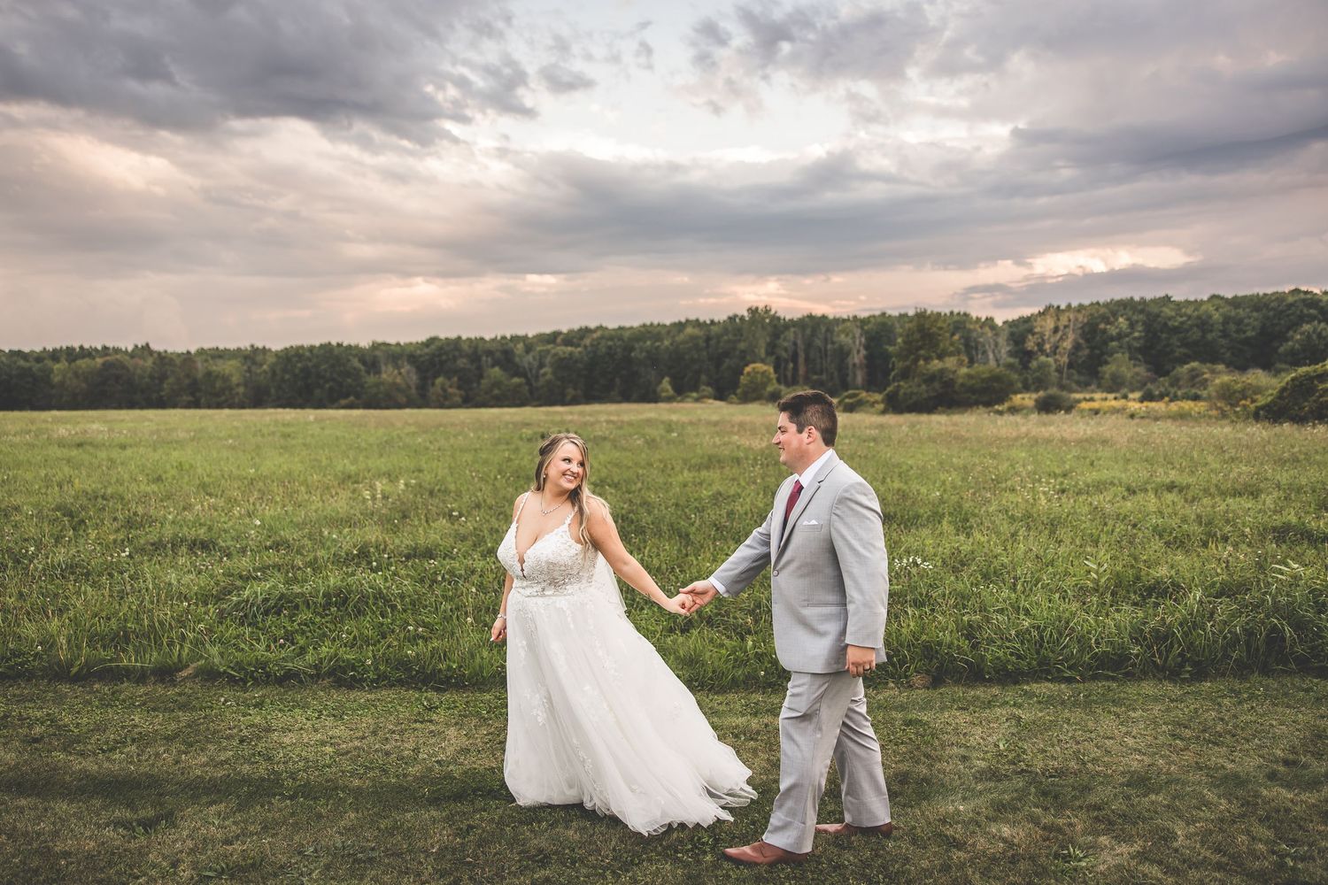 Couple walking hand in hand through grassy field during sunset wedding photoshoot.