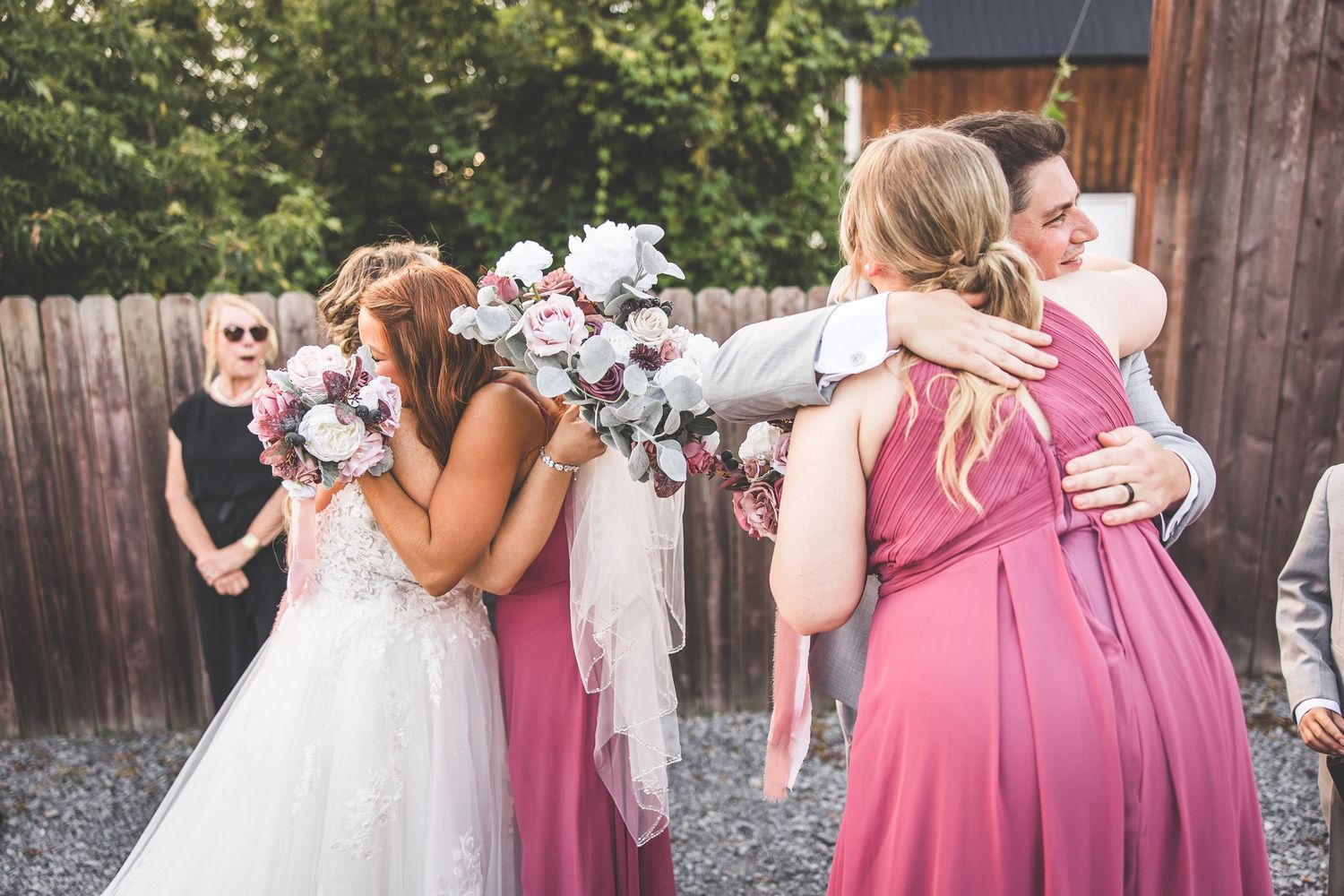 Wedding party members in pink dresses and gray suits pose together outdoors near a rustic barn.