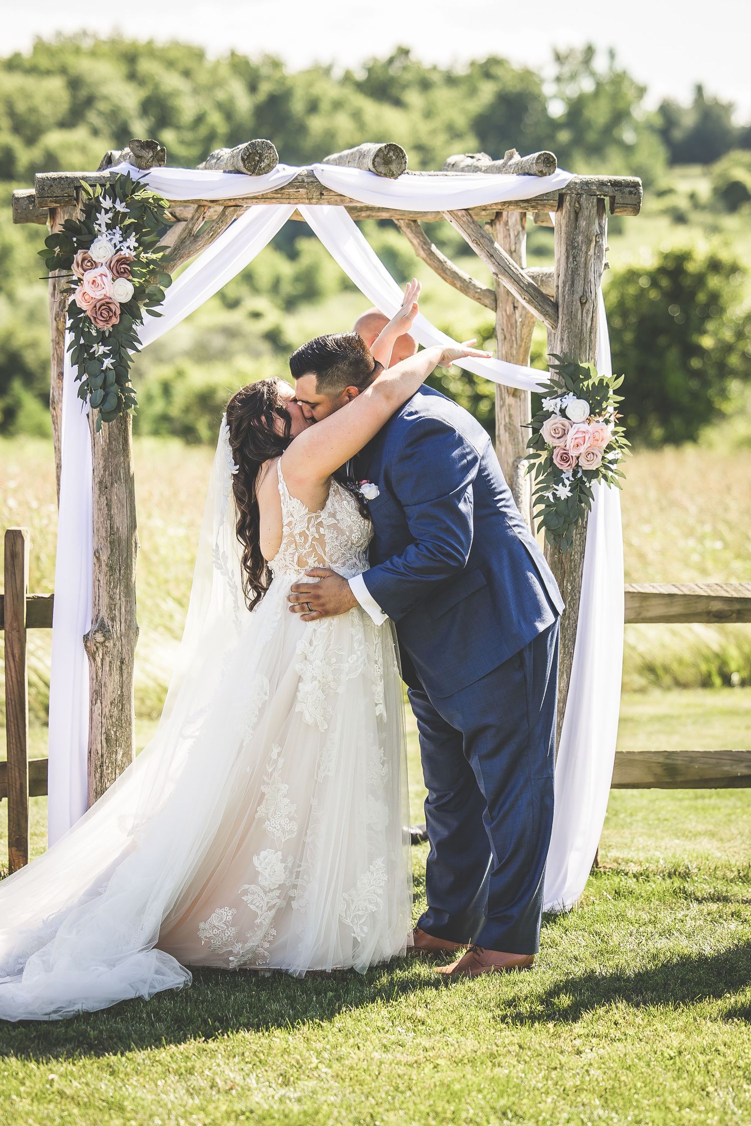 Romantic wedding ceremony moment under rustic wooden arch decorated with white fabric and pink flowers.
