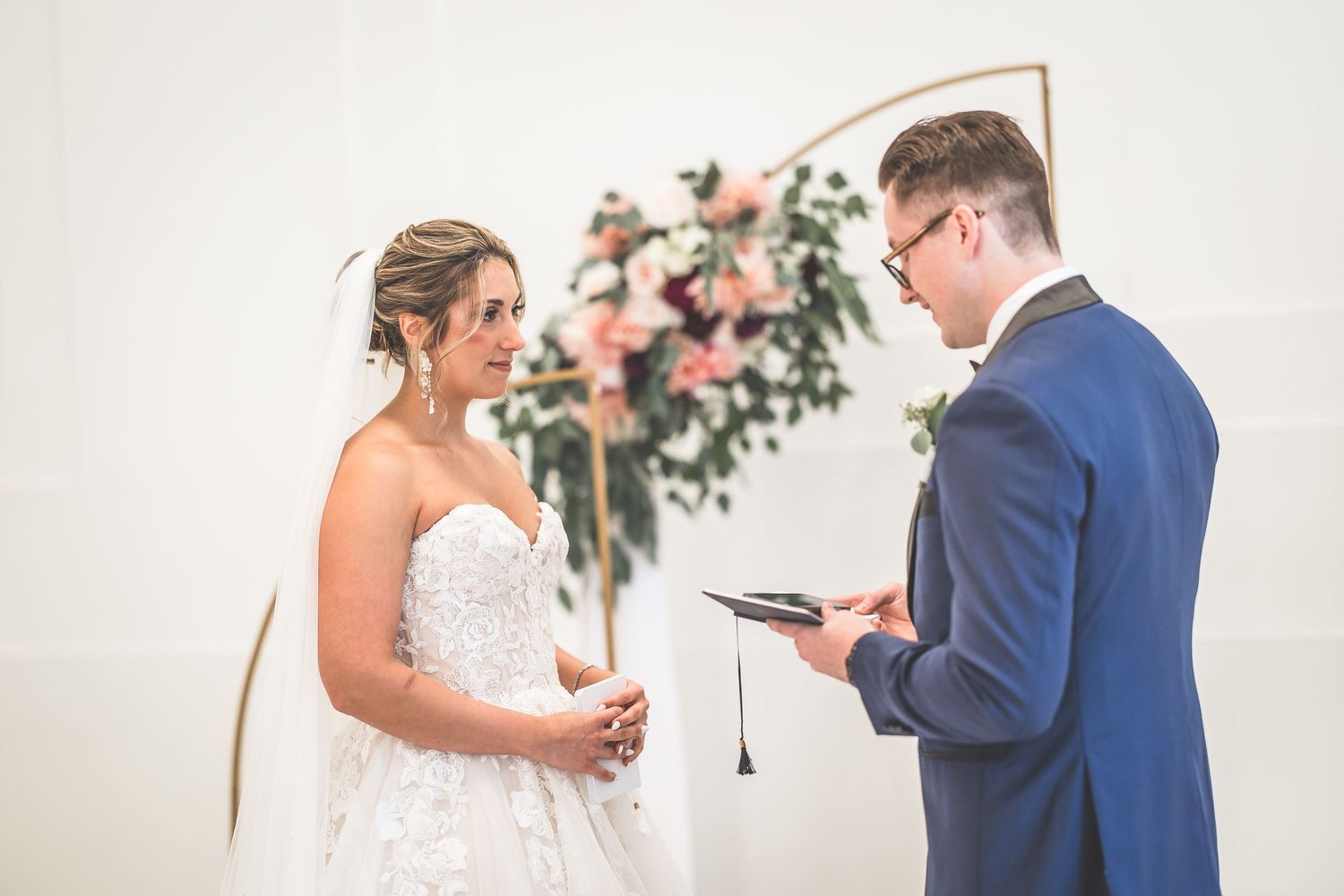A wedding couple exchanges vows in front of a copper arch decorated with pink and white flowers.
