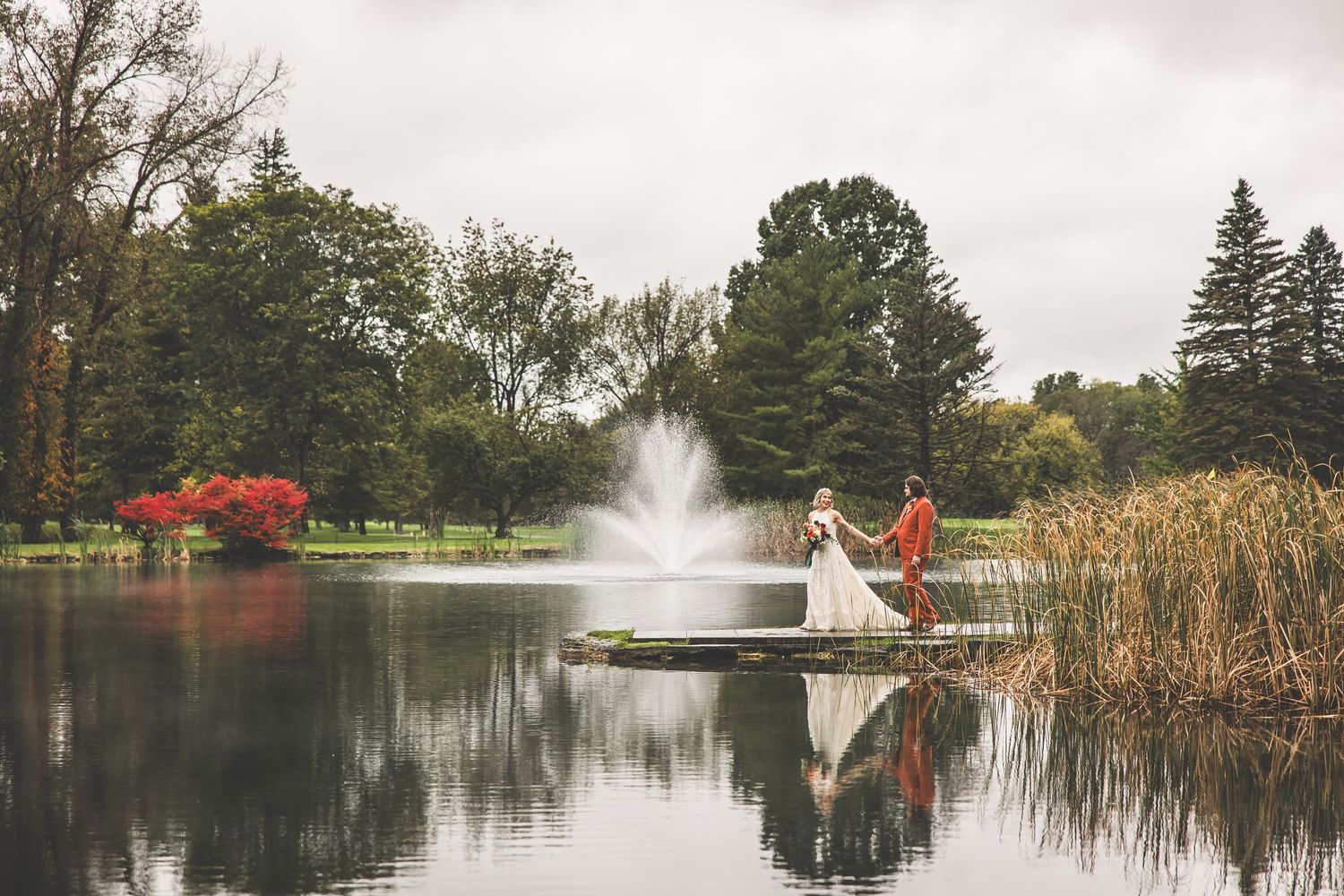 Wedding couple stands on dock with fountain and reflective lake surrounded by autumn landscape.