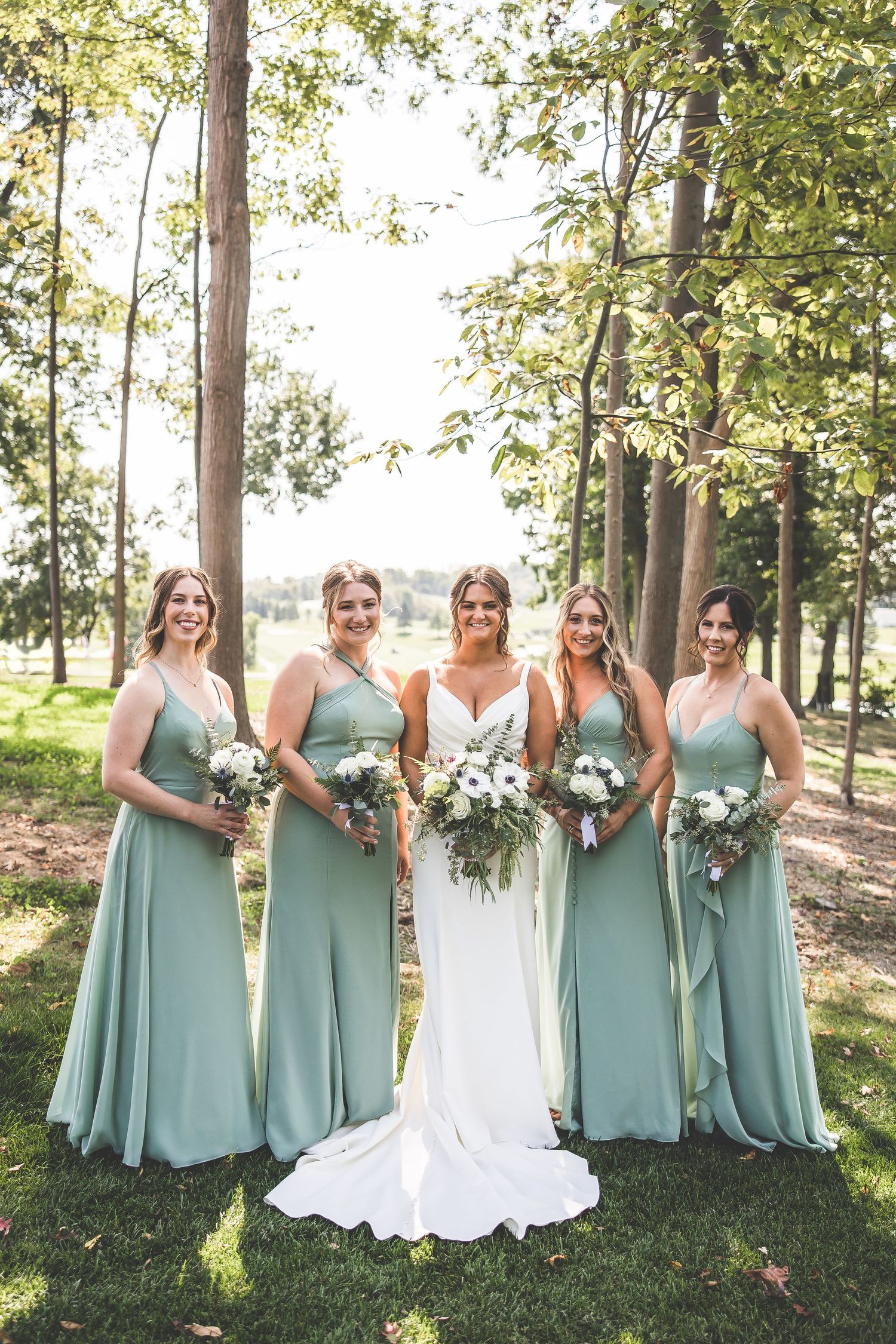 Wedding party in sage green dresses poses with bouquets under tall trees in natural lighting.