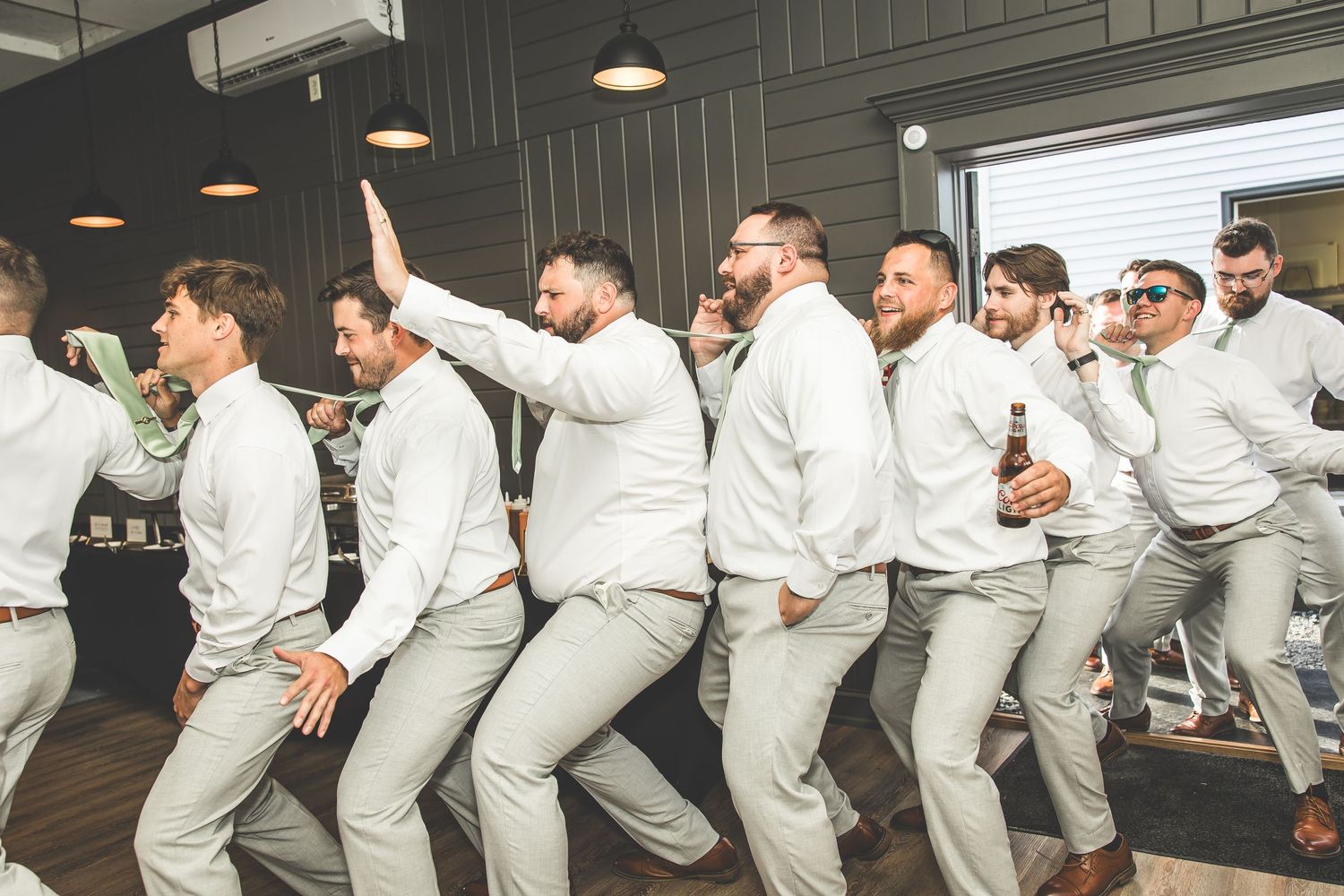 Groomsmen in matching gray suits performing a synchronized dance move indoors.