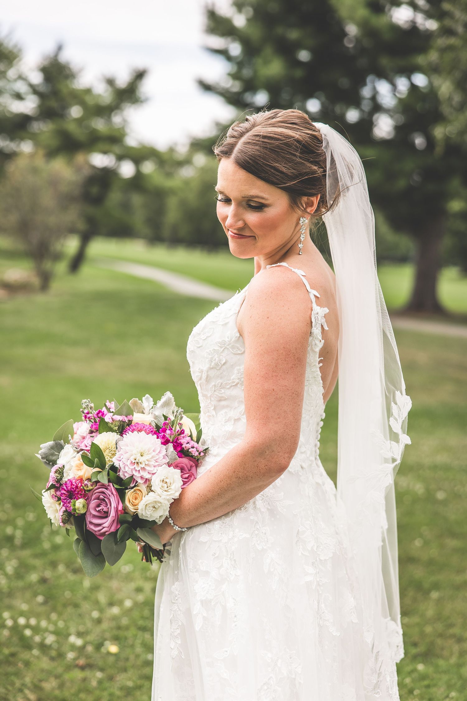 A bride in a detailed lace gown holds a colorful bouquet of purple and pink flowers in a garden setting.