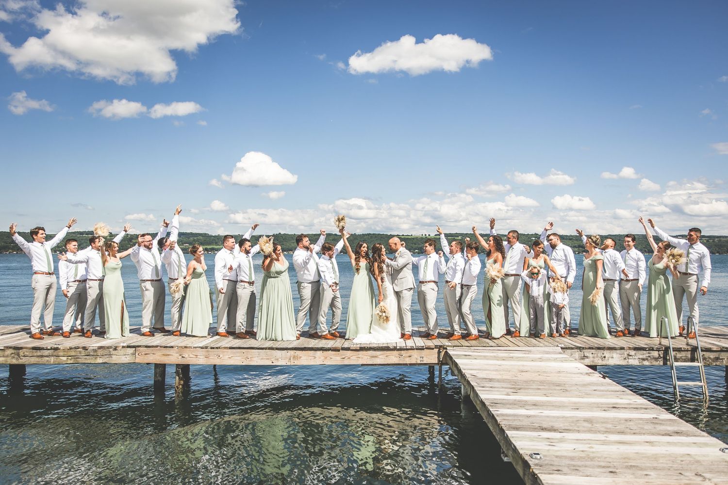 Large wedding party celebrates on lakeside dock wearing coordinated mint and gray attire.