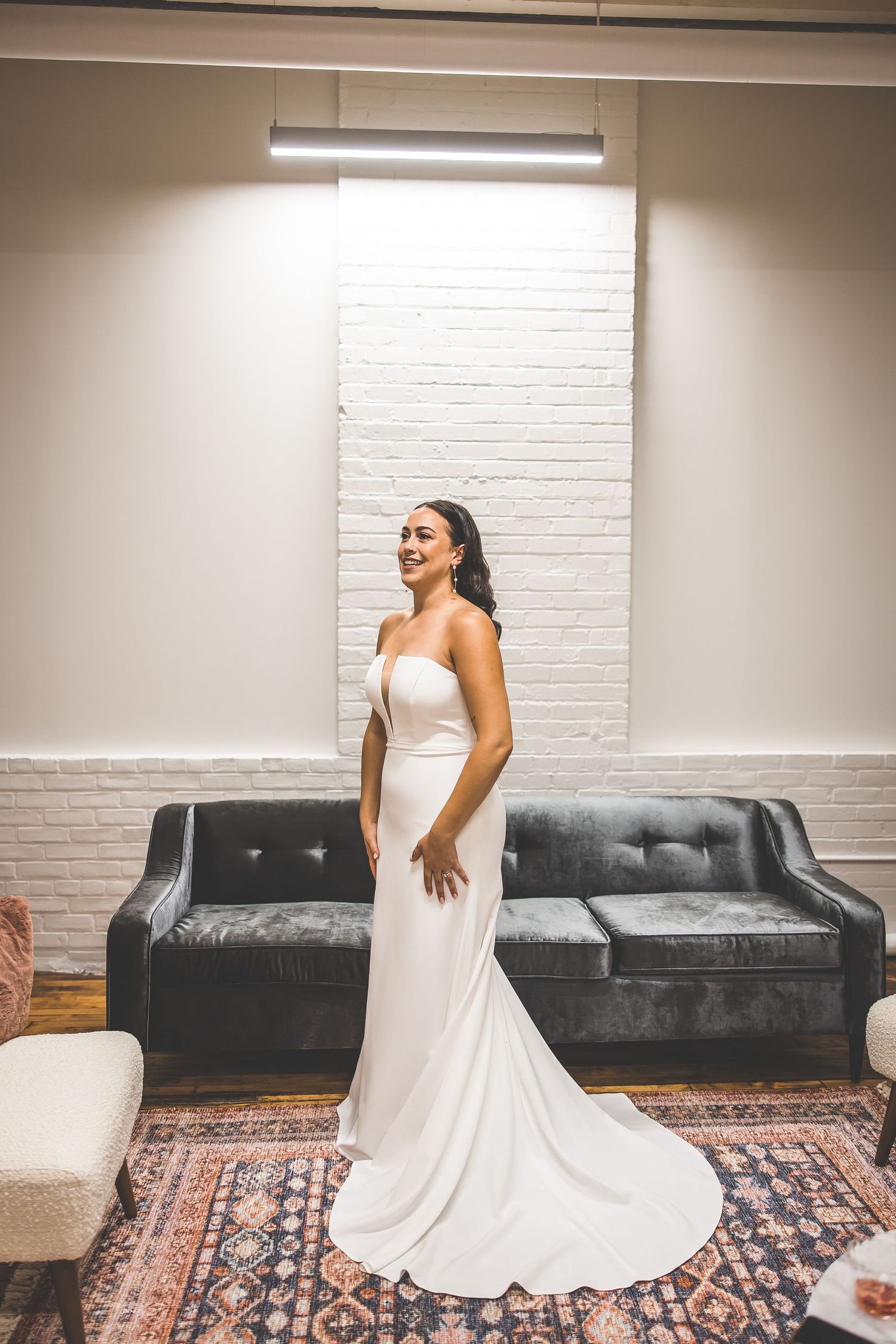 Person in elegant white wedding gown posing on patterned rug near leather couch.