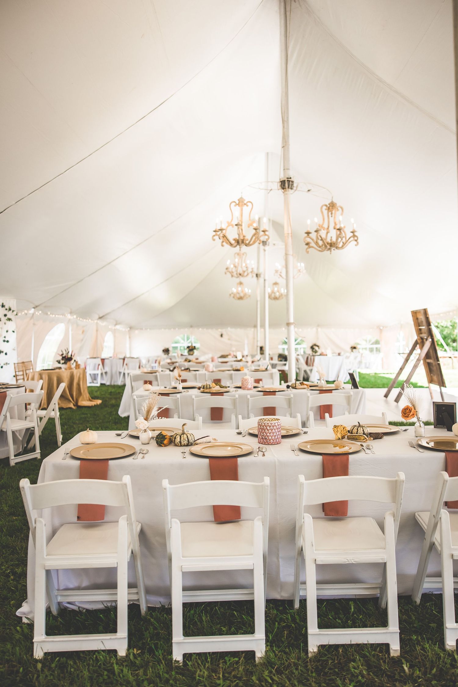 Elegant wedding reception tent with chandeliers and white folding chairs at round tables.