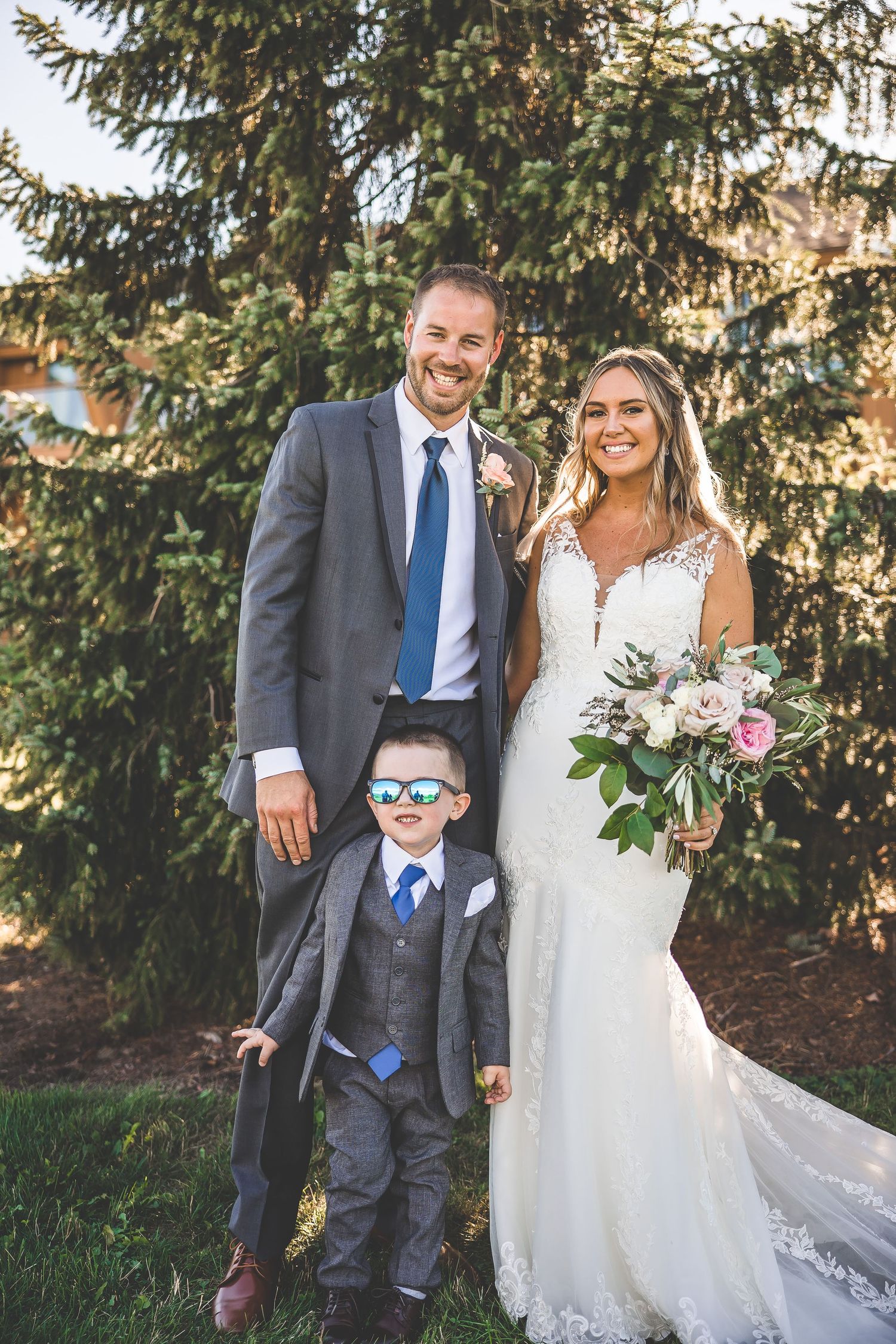 Outdoor wedding portrait with a young ring bearer in sunglasses against evergreen trees.