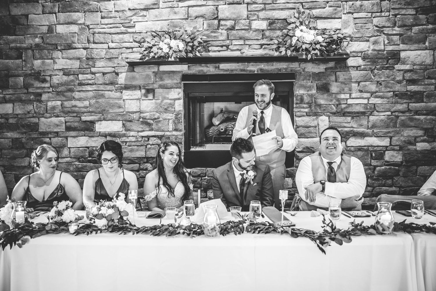 Wide shot of wedding party seated at head table against stone wall background.
