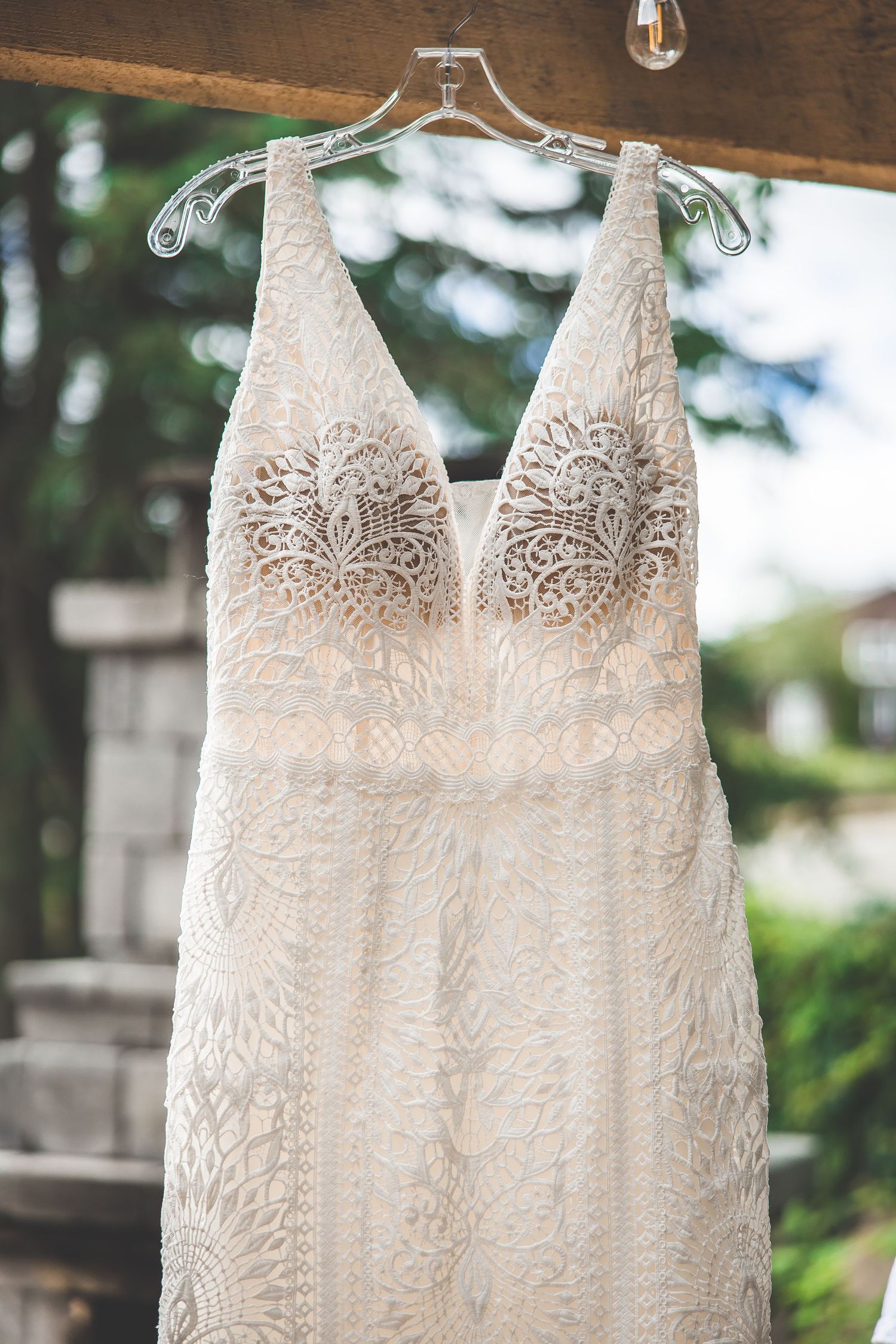 A delicate lace wedding dress hangs on display with string lights in background.