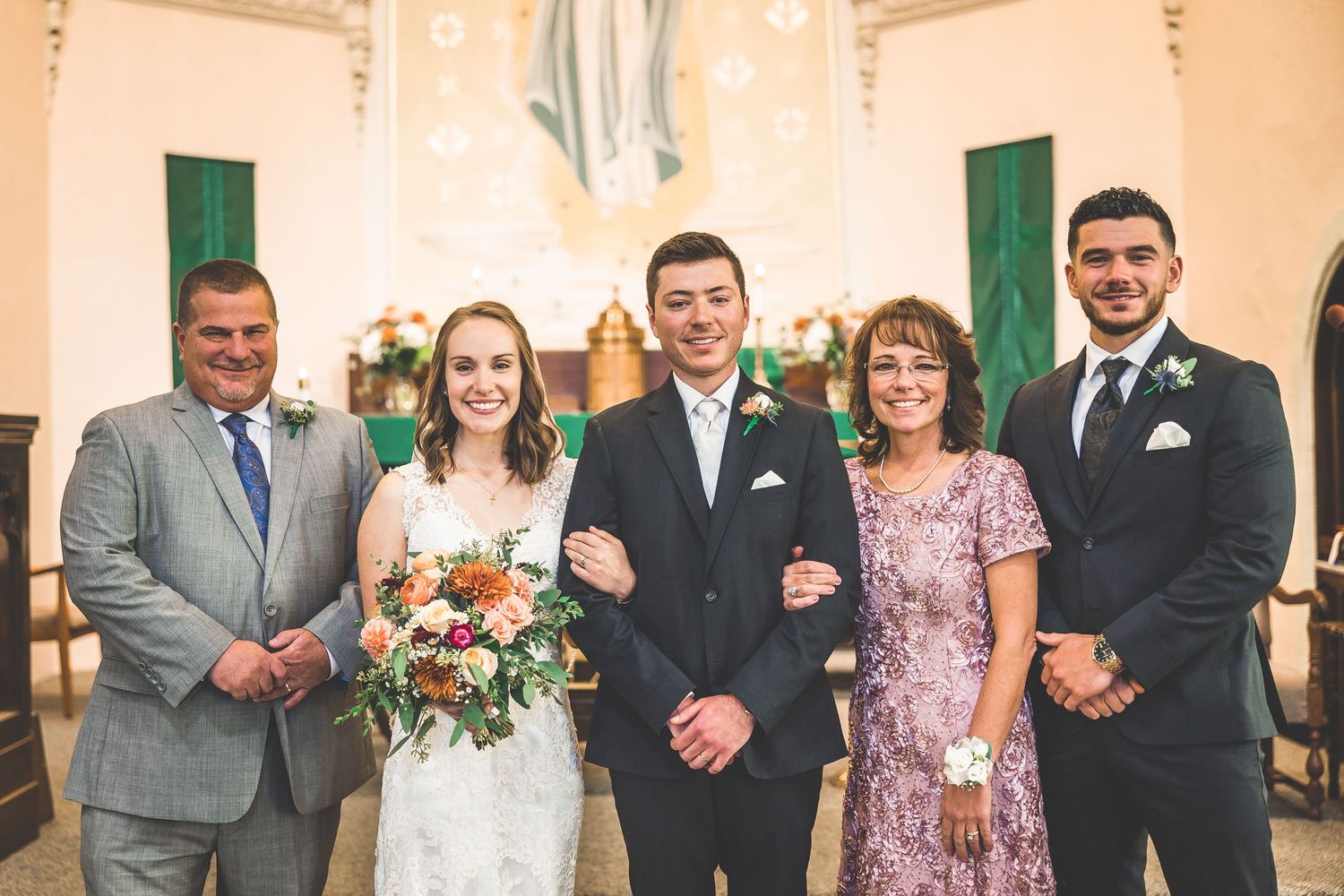 Wedding party poses together in formal attire with burgundy bridesmaid dresses and black suits in church.
