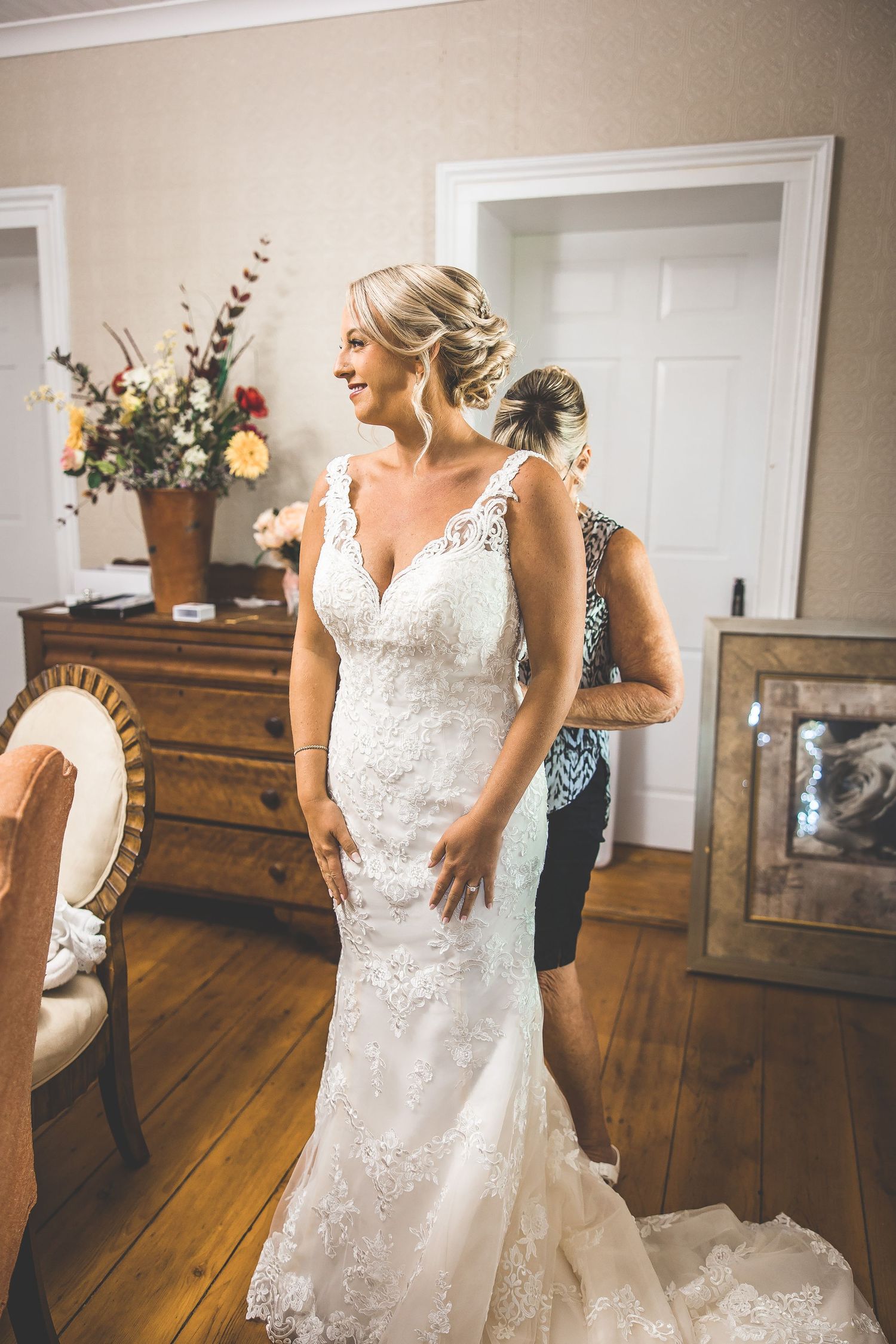 Bride in lace wedding gown standing in elegant interior with wooden floors and vintage furniture.