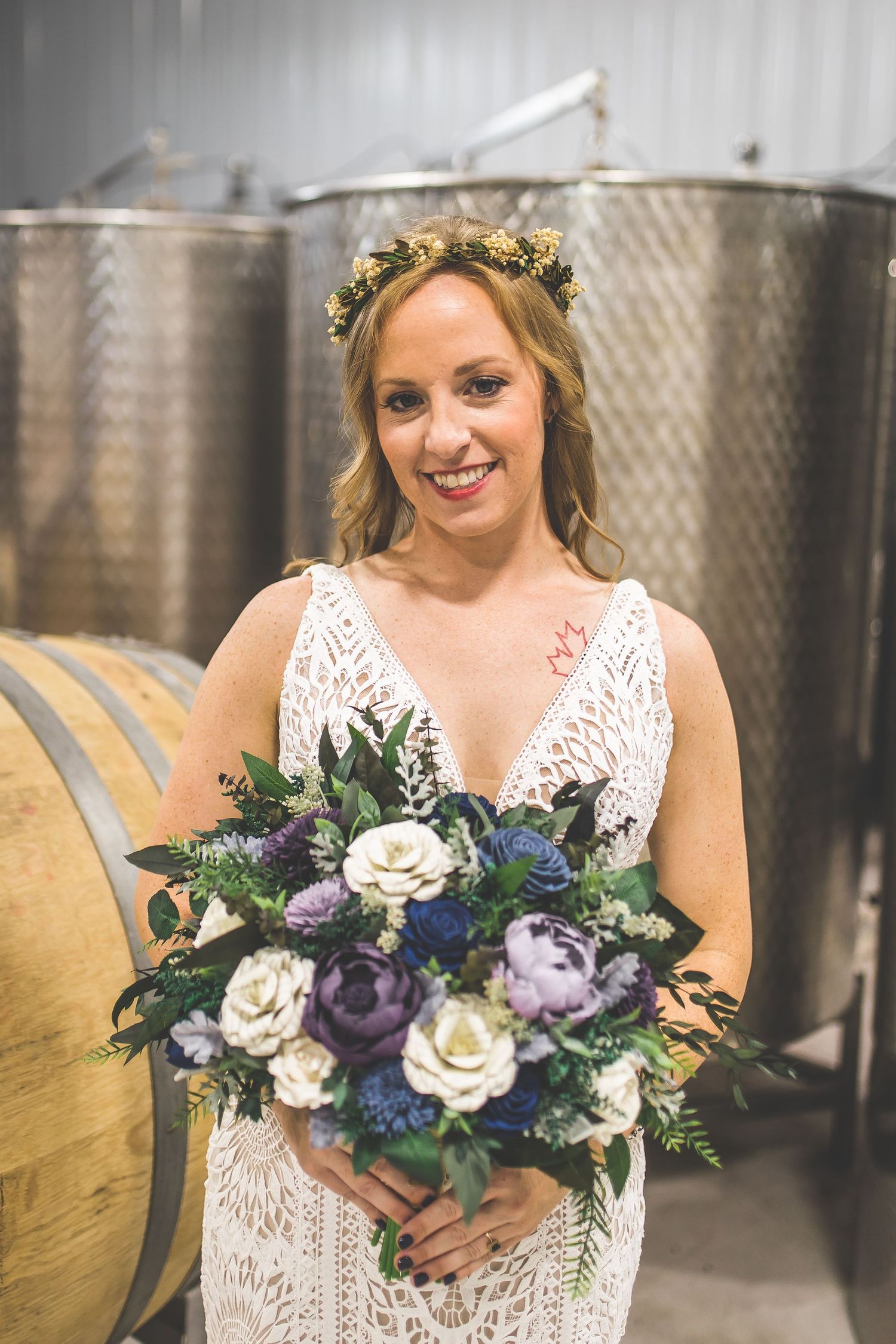 Wedding party poses with floral bouquets featuring blue and purple blooms against metal wine tanks.