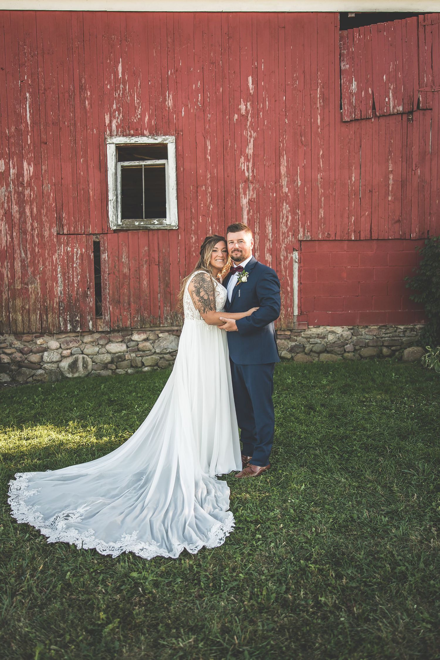 A couple in wedding attire poses against a rustic red barn with a flowing white dress train on grass.