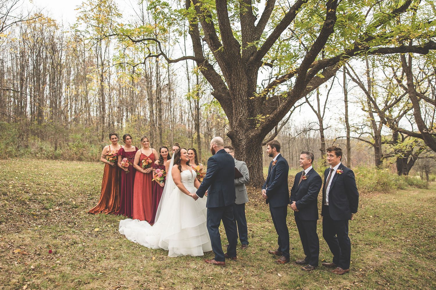 Wedding ceremony in progress under large trees with burgundy-clad bridesmaids and navy-suited groomsmen.