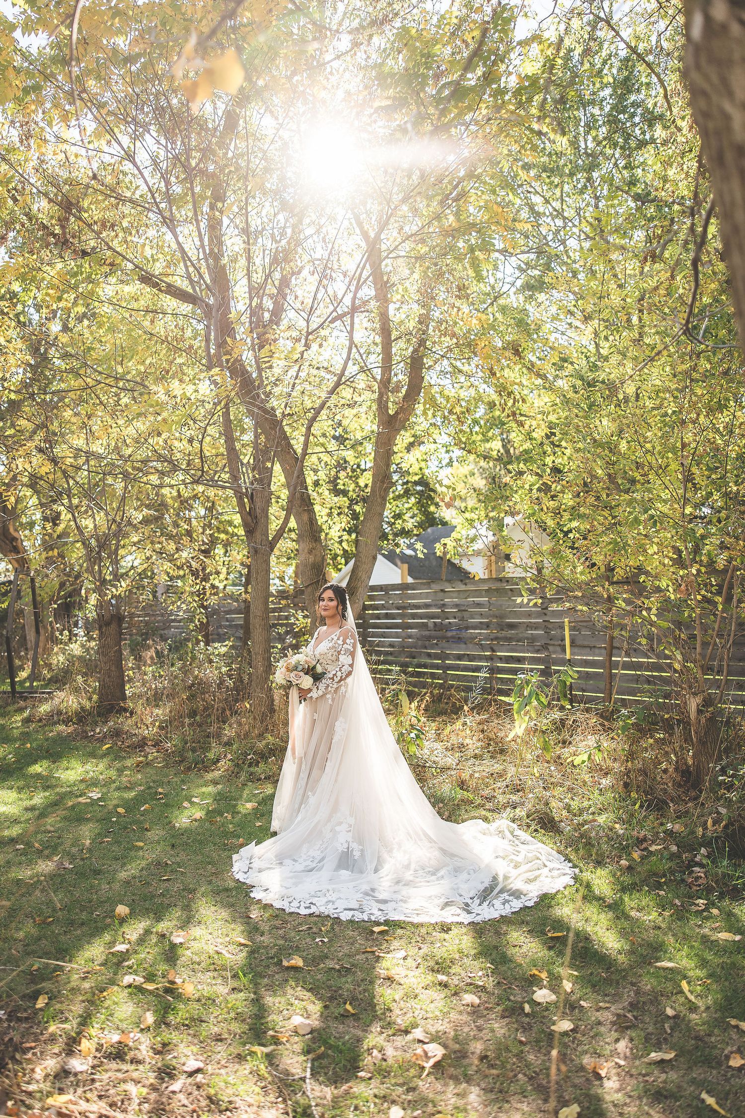 Elegant white wedding dress with long train photographed in a sunlit wooded outdoor setting.