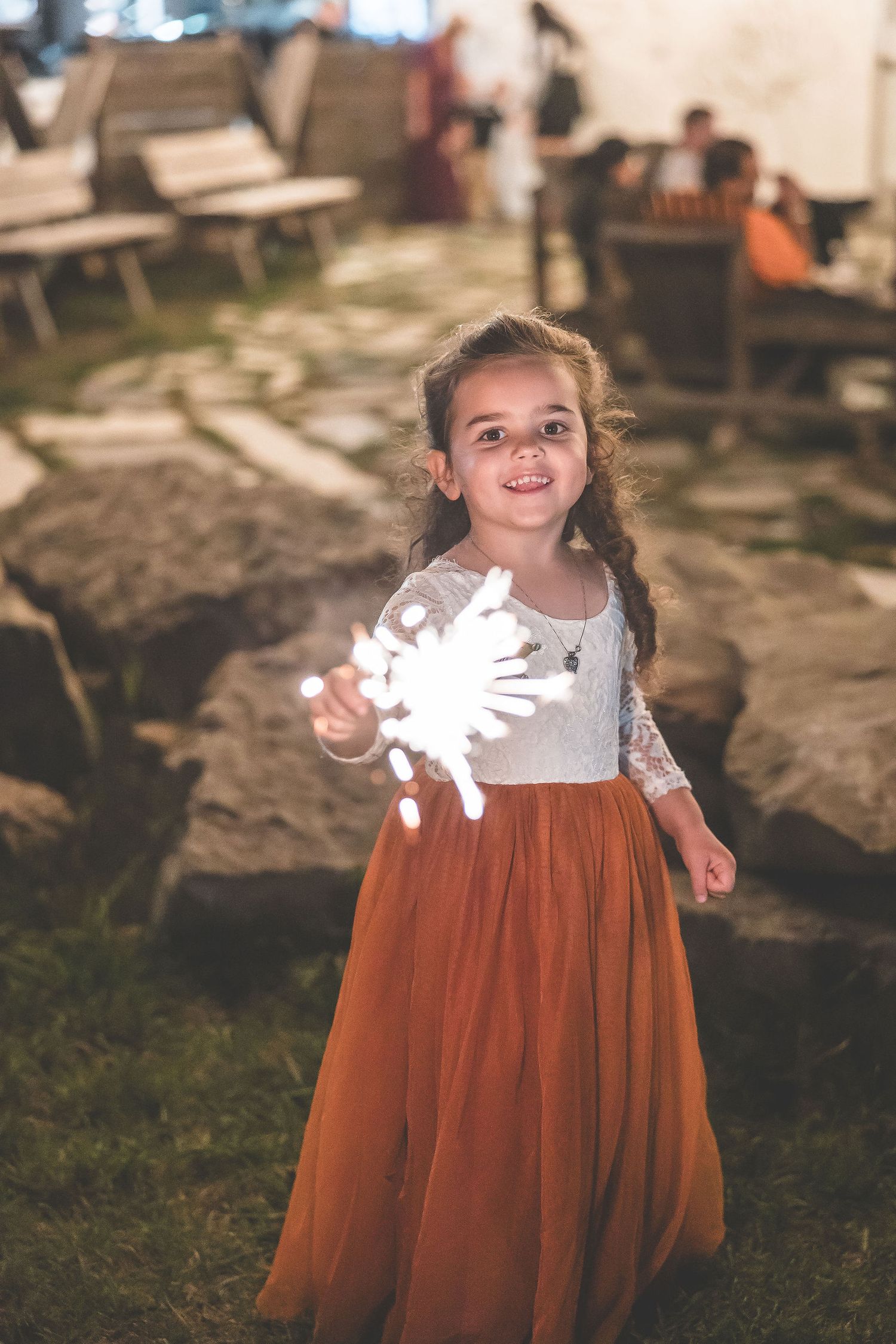 Young girl in a rust-colored dress twirls while holding a sparkler outdoors.