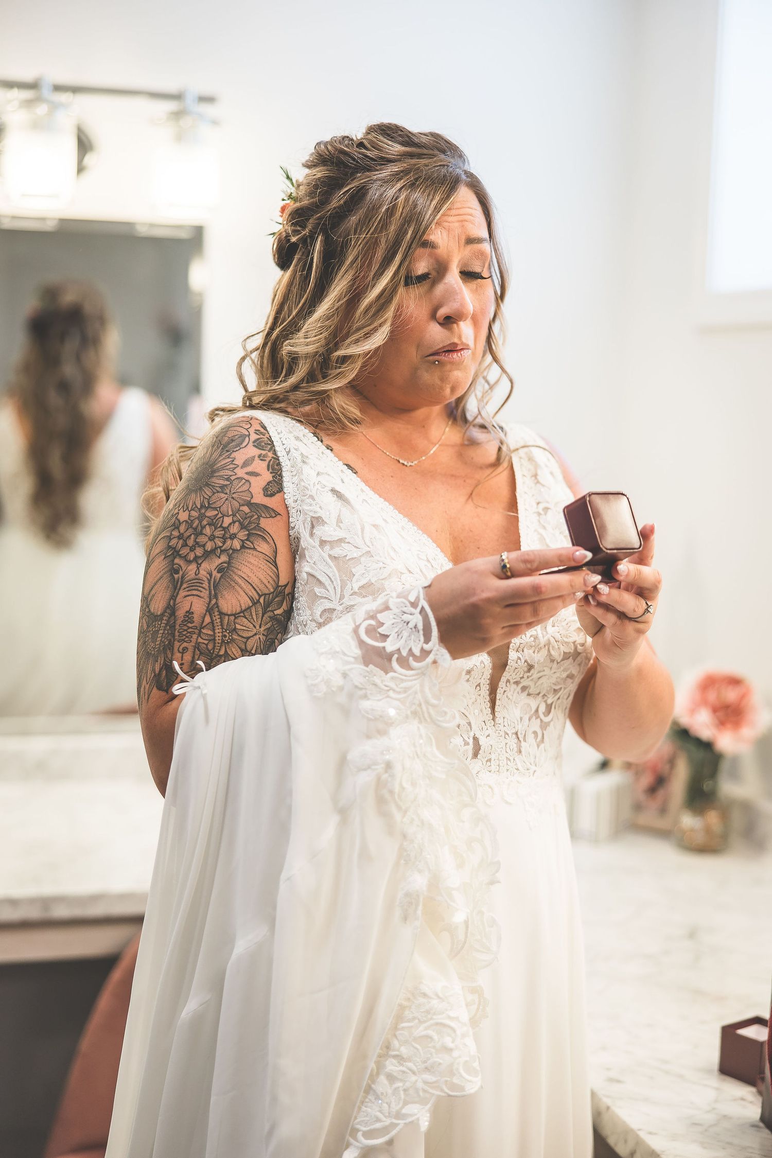 Bride in white lace dress holds her gown while getting ready in a bright dressing room.