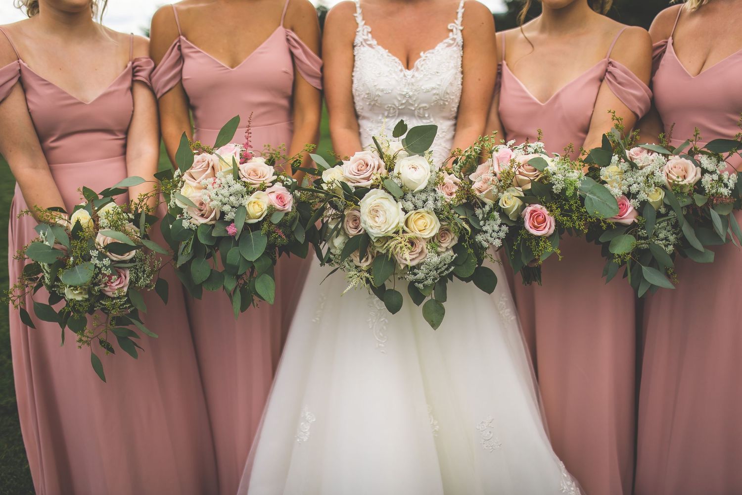 Close-up of bridesmaids in pink dresses holding elegant floral bouquets at outdoor wedding.