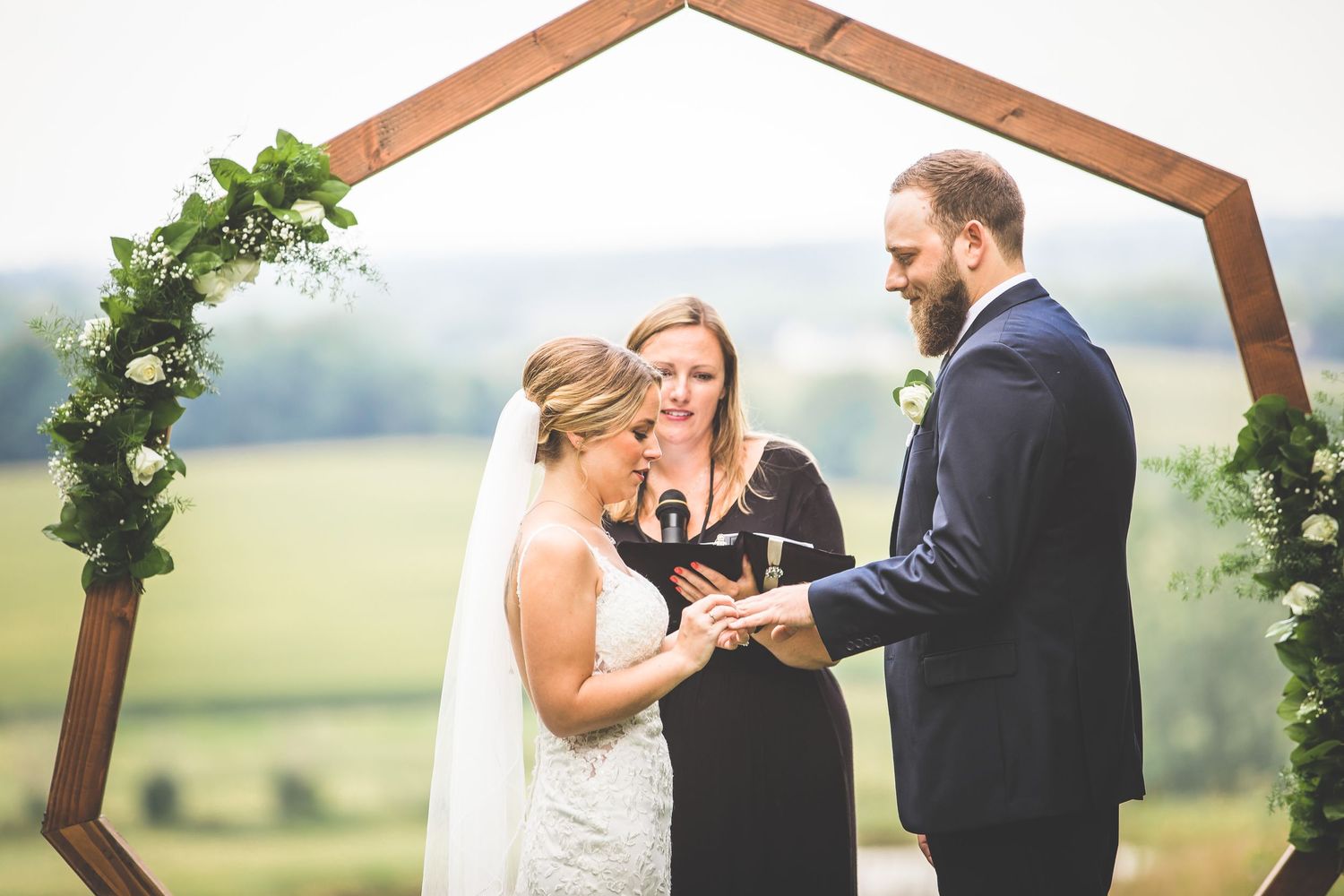 Intimate wedding ceremony moment under wooden arch decorated with greenery against countryside backdrop.