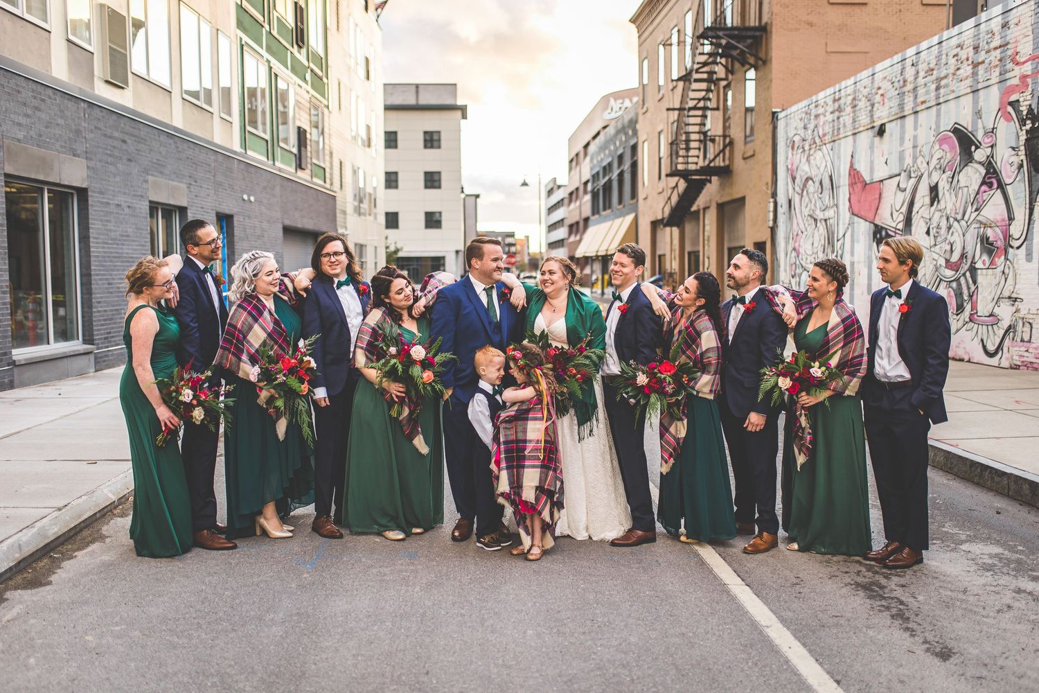 Wedding party members celebrate on urban street with graffiti wall backdrop.