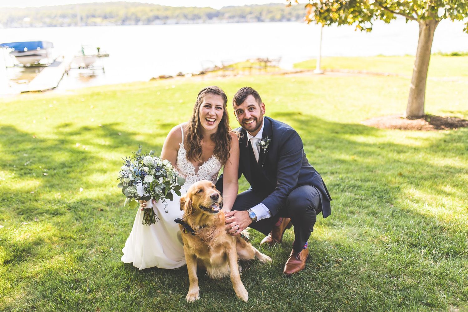 Wedding couple poses with their golden retriever on a sunny lakeside lawn.