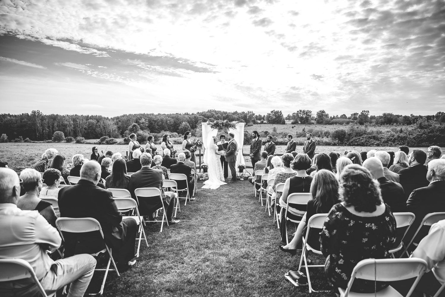 Black and white panoramic view of wedding guests seated in rows of white chairs at an outdoor ceremony.