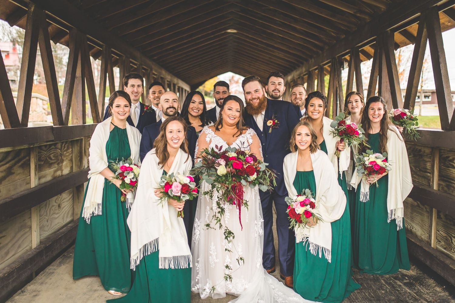 Bridal party celebrates in a rustic covered bridge with bridesmaids in emerald dresses.