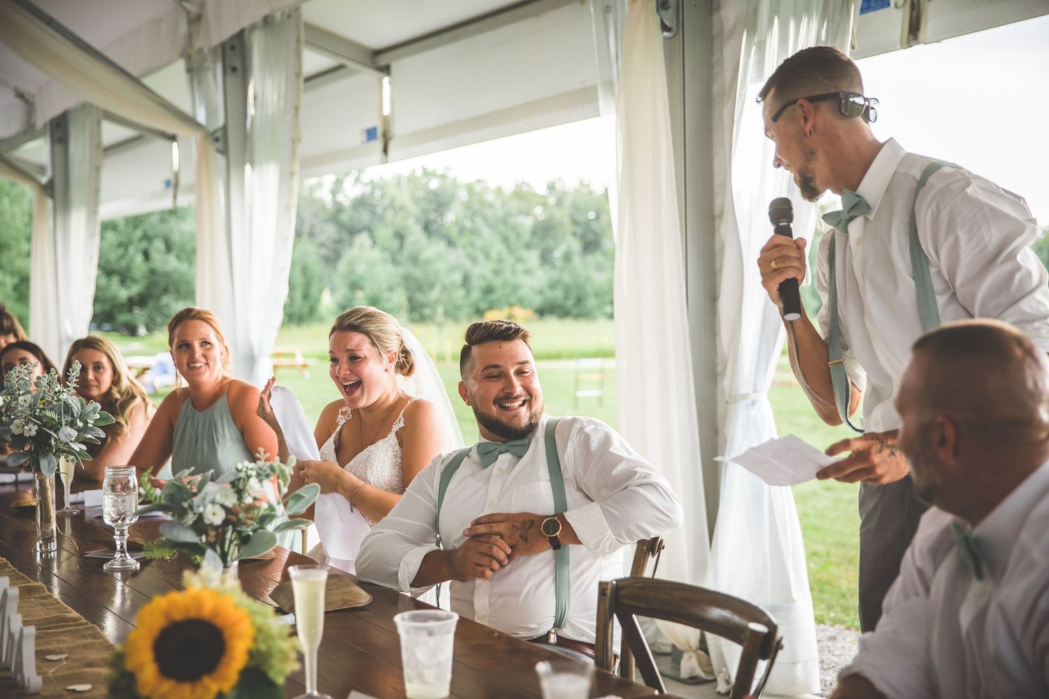 Wedding guests and party members laugh during speeches at a sunflower-decorated reception.