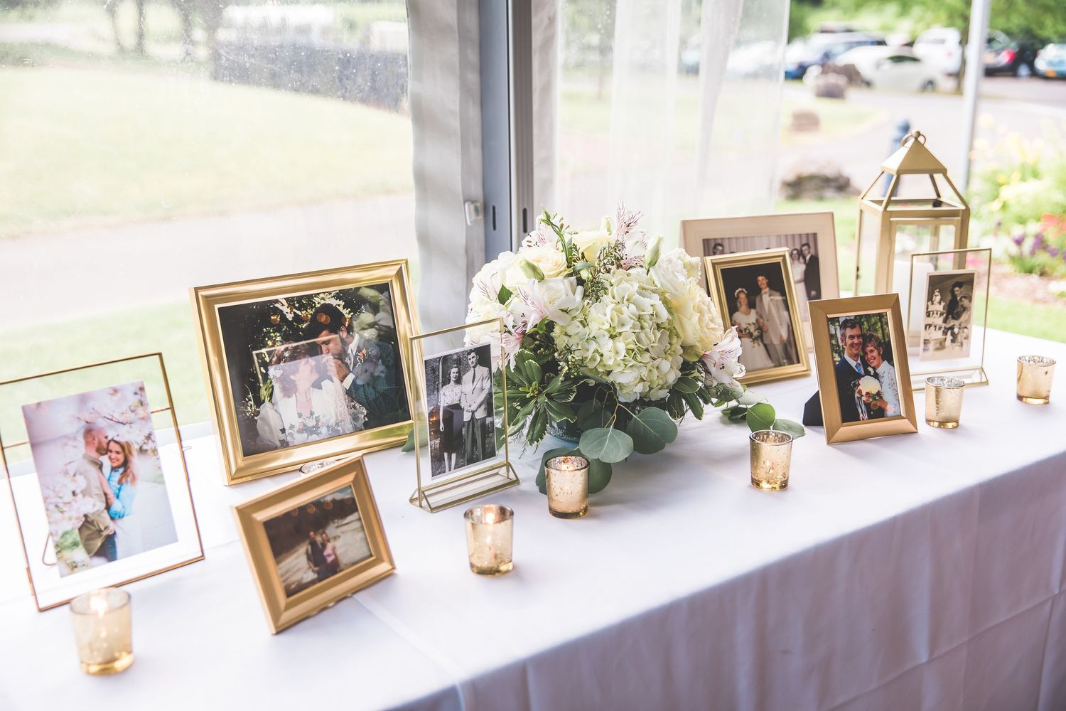 Wedding photo display table with framed pictures and decorative elements near large windows.